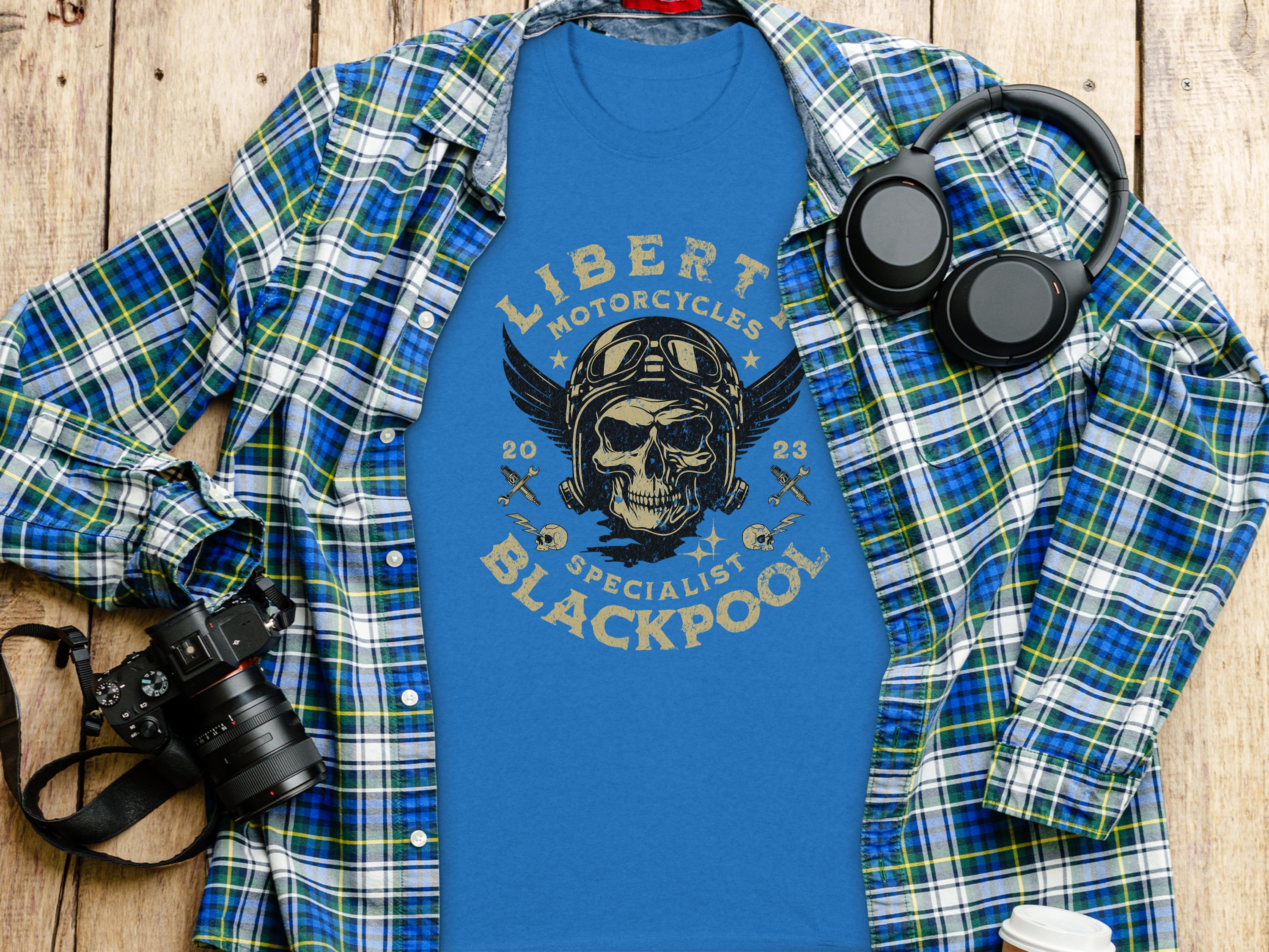 A Garment Graphics blue t-shirt with a skull and motorcycle graphic, featuring Liberty Motorcycles Blackpool, lies on wood, accompanied by a blue plaid shirt, headphones, a coffee cup, and a camera—perfect for motorcycle enthusiasts.