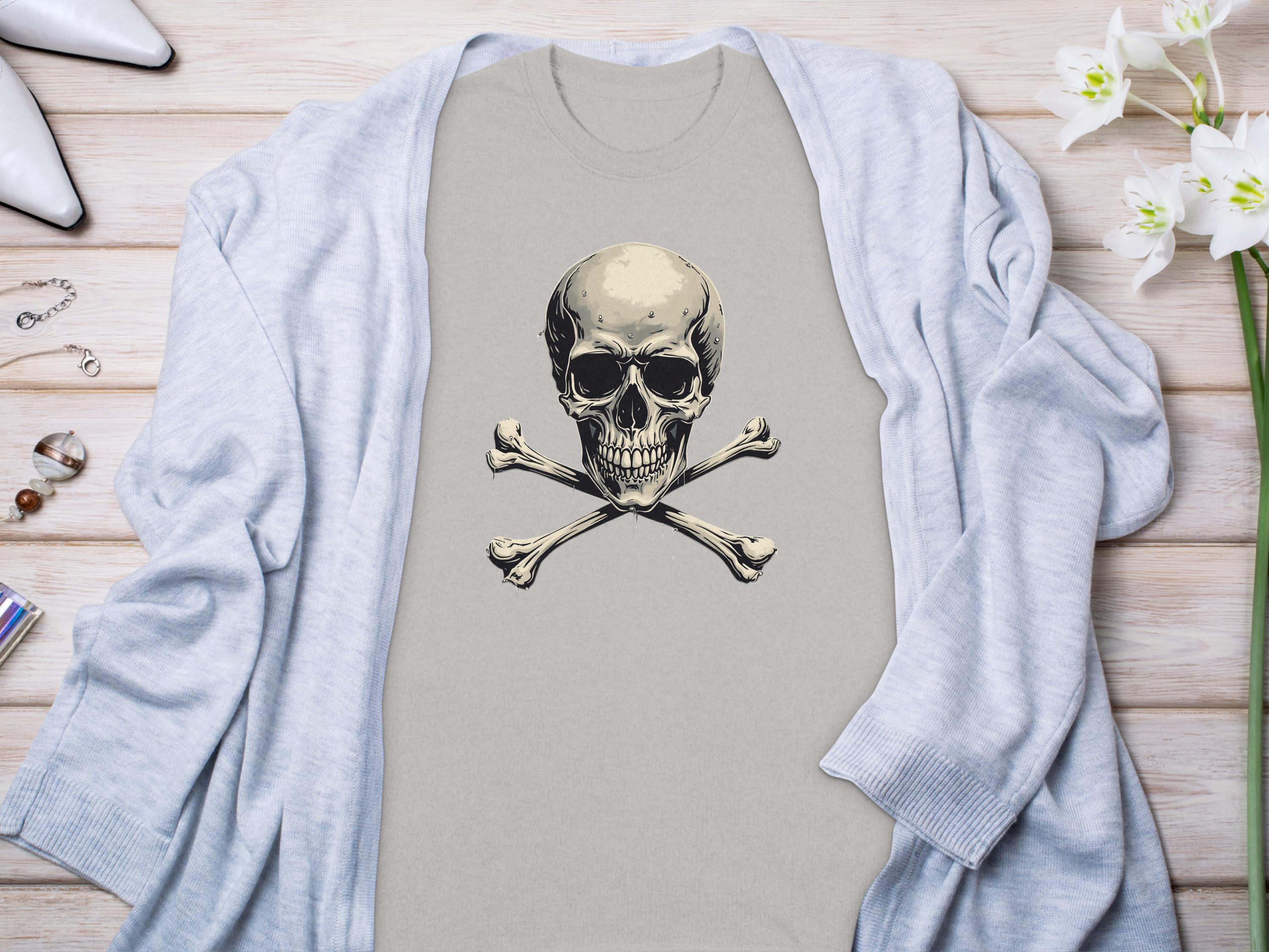 A beige skull and crossbones graphic t-shirt by Garment Graphics is styled on a light wooden surface. A gray cardigan drapes over it, enhancing the chic look. White high-heeled shoes, a bracelet, ring, and white flowers complete the fashionable ensemble.