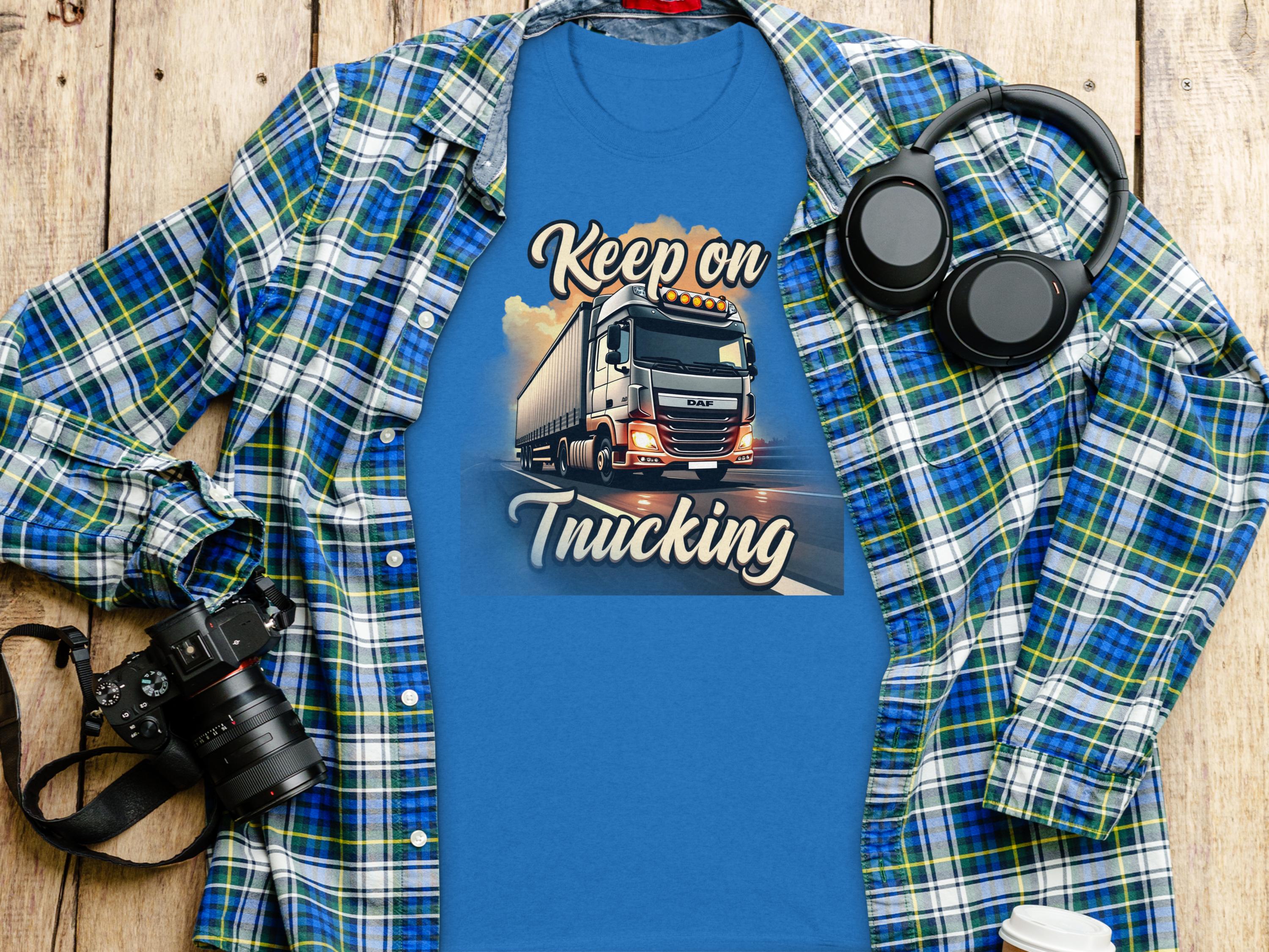 Garment Graphics offers a blue T-shirt with a semi-truck graphic and Keep on Trucking text, ideal for truck enthusiasts. Its styled with a plaid shirt, headphones, camera, and coffee cup on a wooden surface.