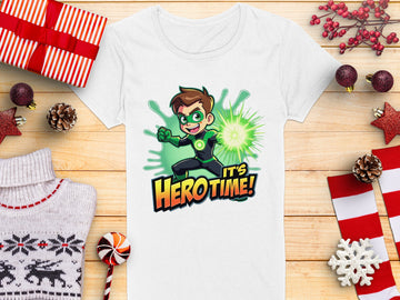 Garment Graphics white t-shirt showcases young heroes, including a brown-haired cartoon in green superhero gear with a vibrant energy aura. The phrase Its Hero Time! graces the print. Displayed on wood, its surrounded by festive décor, gift boxes, and pinecones—a colorful wardrobe staple!.