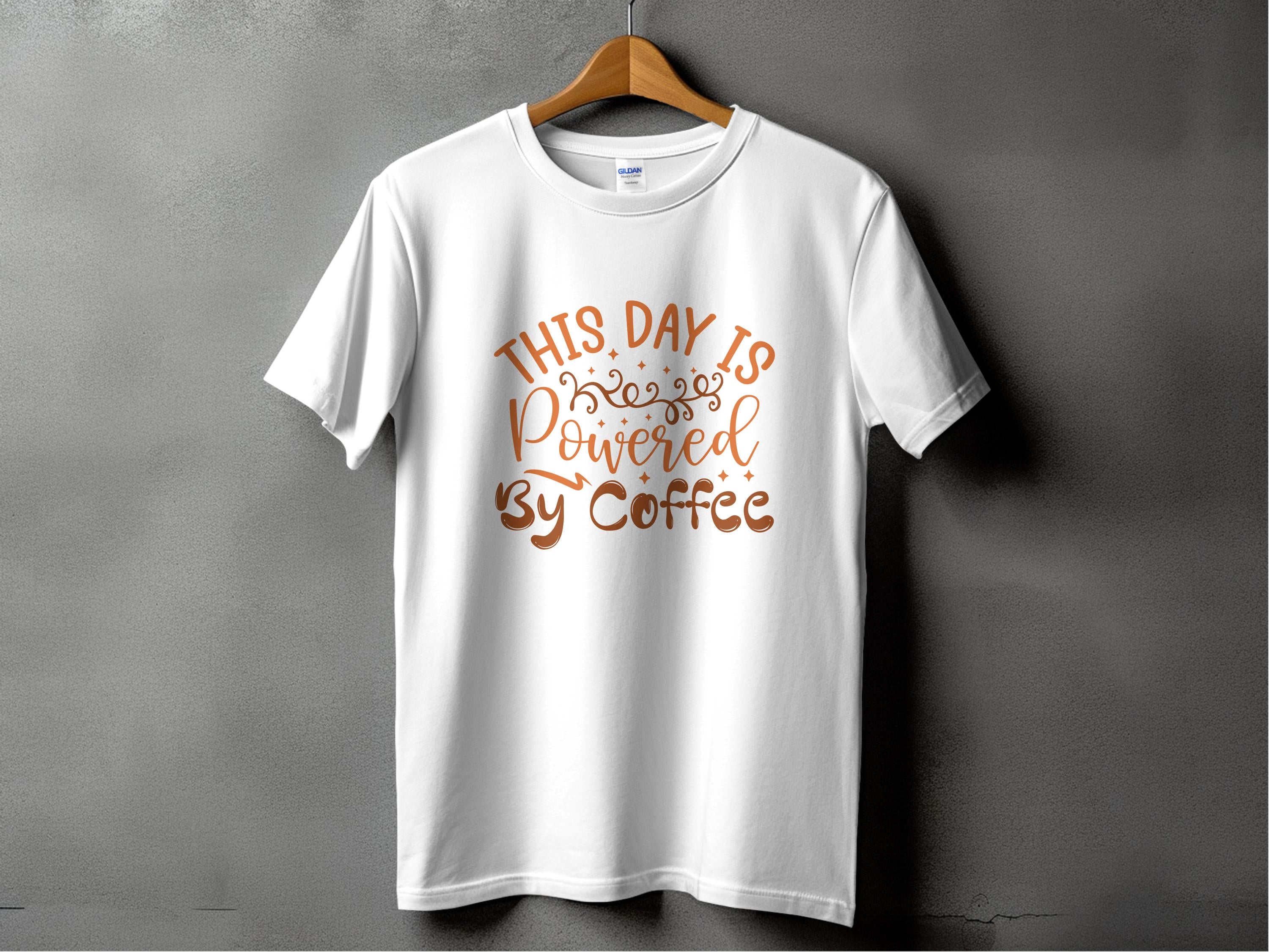 A white classic fit t-shirt from Garment Graphics hangs against a gray backdrop, displaying This Day is Powered by Coffee in bold brown cursive with decorative swirls—ideal for coffee lovers and positivity enthusiasts.