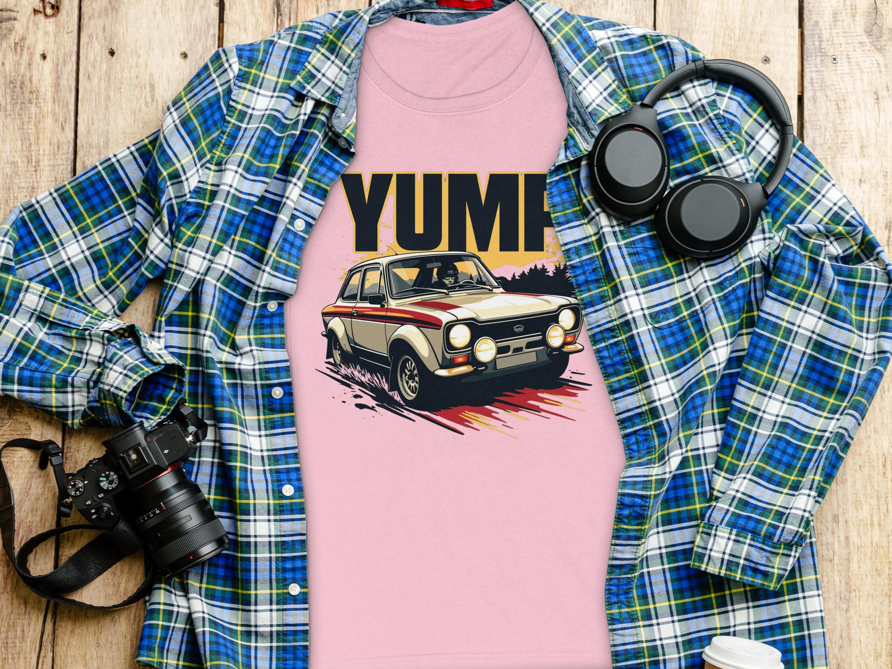 A pink Garment Graphics tee featuring a vintage car graphic and YUMP lies on wood, layered with a blue plaid shirt. Nearby are a camera, headphones, and a white coffee cup.