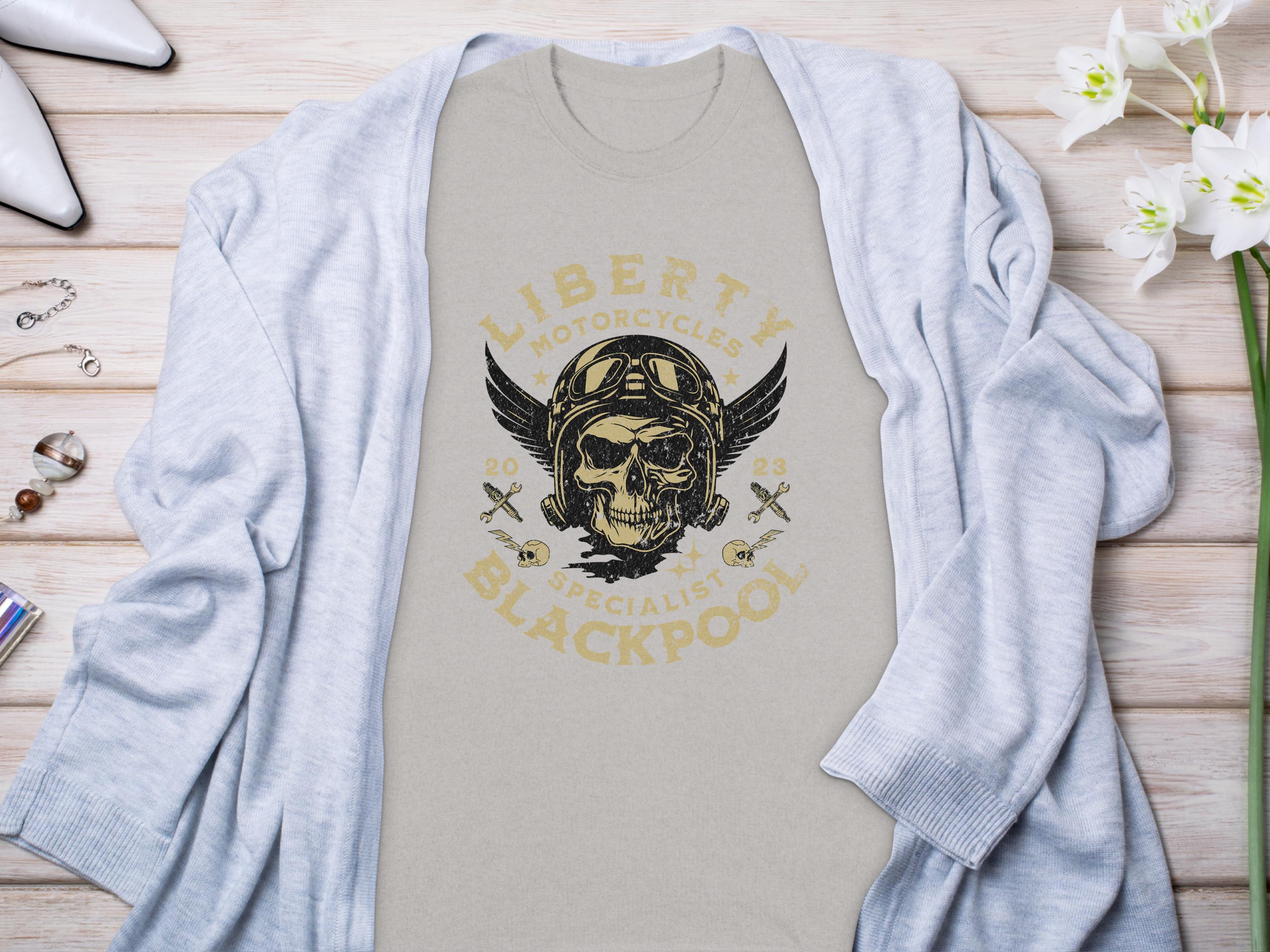 A light gray T-shirt by Garment Graphics features a vintage design of a winged skull in a helmet and goggles, surrounded by Liberty Motorcycles Specialist Blackpool 2023. Perfect for motorcycle enthusiasts, its styled on a wooden surface with a gray cardigan, flowers, and accessories.
