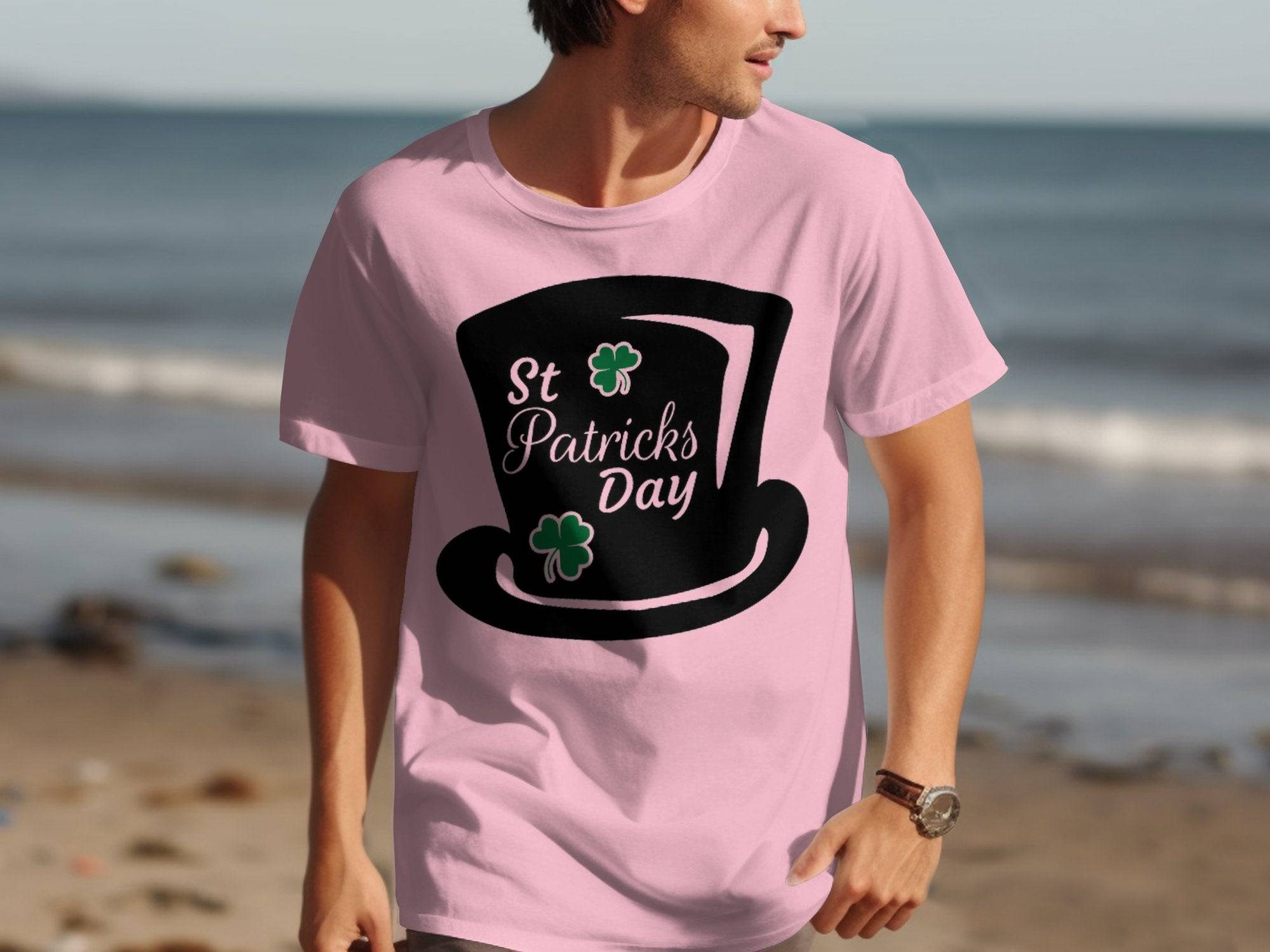A person walks on the beach wearing a Garment Graphics pink T-shirt featuring a large black top hat with the text St Patricks Day, adorned with two green shamrocks. The serene ocean backdrop enhances this festive attire.