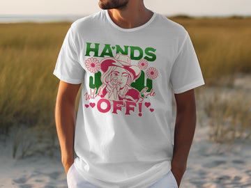 A person sports a Garment Graphics classic fit white T-shirt, showcasing a cartoon figure in a cowboy hat, surrounded by cacti and flowers with the text HANDS Wild OFF! in green and pink, set against a grassy landscape and sandy backdrop.