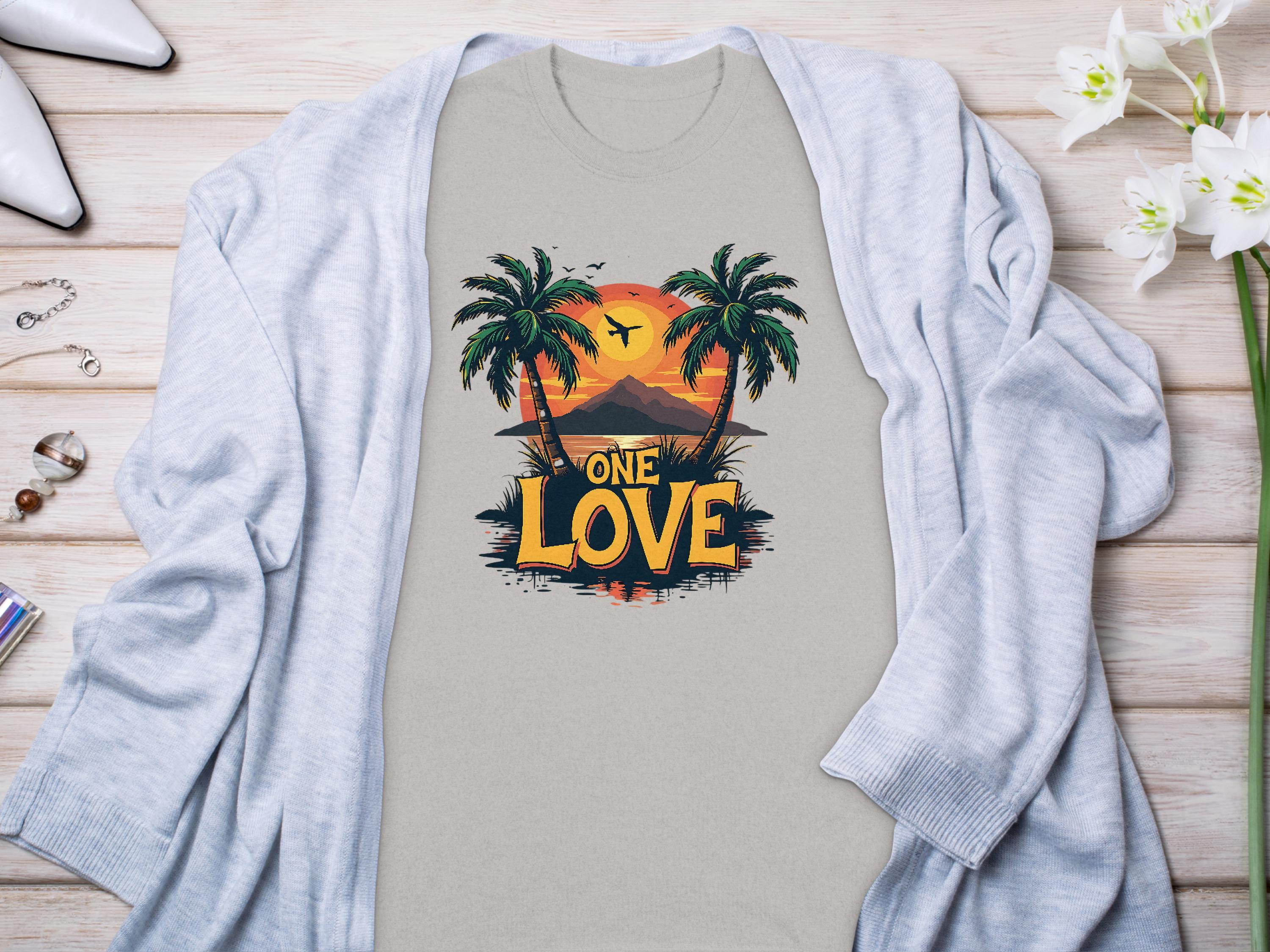 On a wooden surface, the Garment Graphics T-shirt with two palm trees, a tropical sunset, and ONE LOVE in bold is framed by a light gray cardigan. Perfect for beach lovers, its styled with white high heels, jewelry, and white orchids for a complete look.