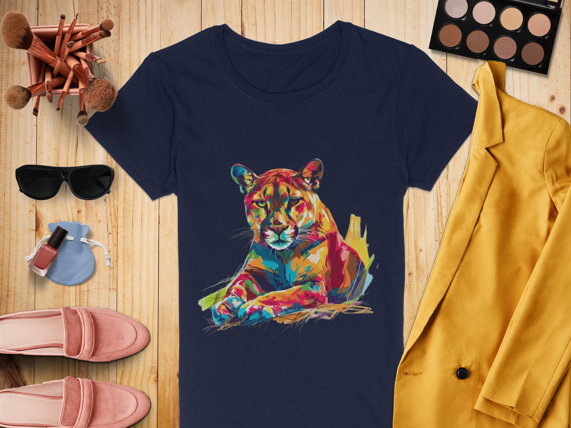 A flat lay image featuring Garment Graphics navy T-shirt with a colorful puma design, complemented by a yellow jacket, makeup palette with brushes, sunglasses, pink shoes, nail polish, and a small blue purse on a wooden background.