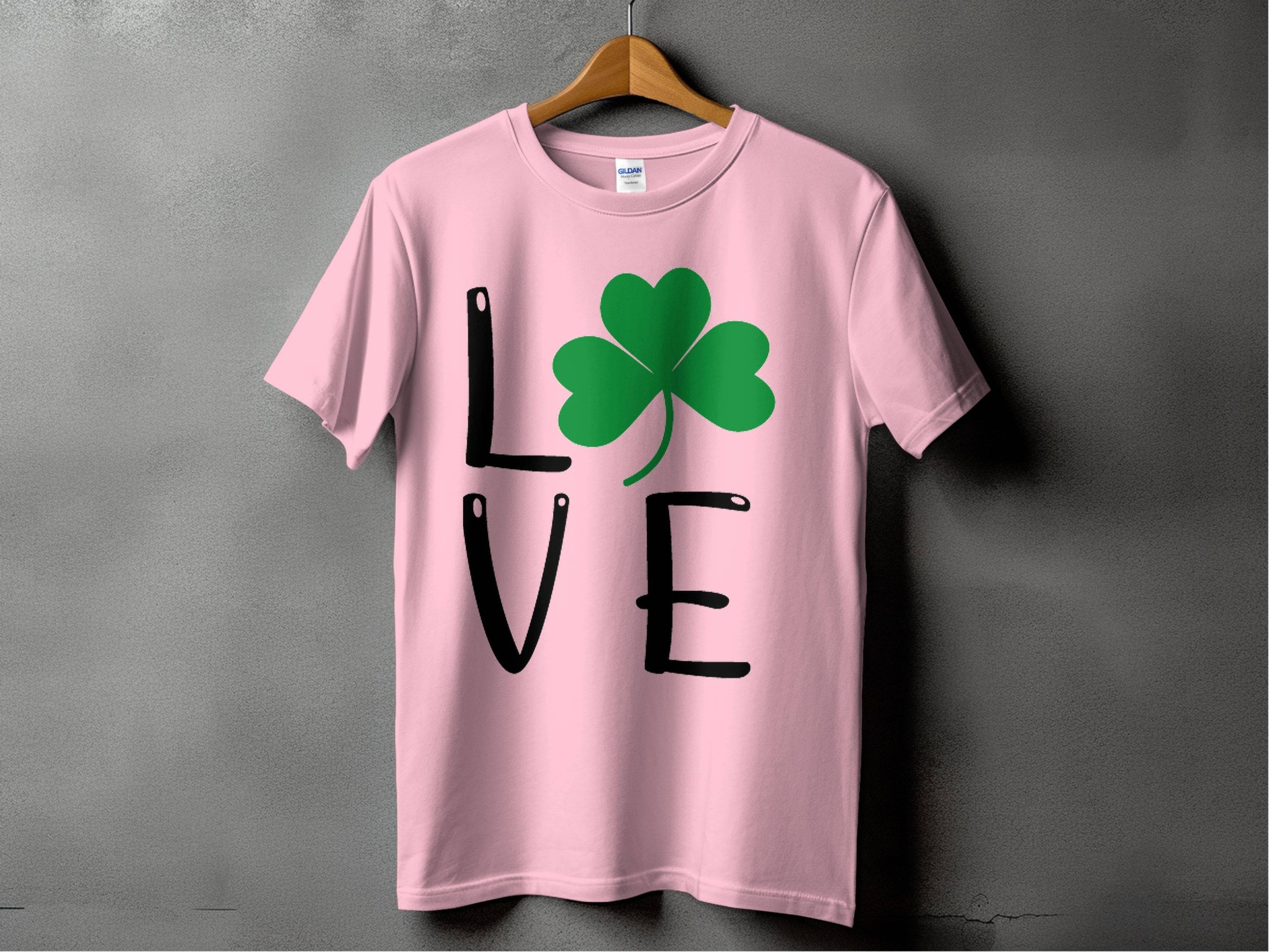 A pink T-shirt by Garment Graphics hangs on a wooden hanger, showcasing Irish spirit with L💚VE, where a green shamrock replaces the O. This charming St. Patricks Day shirt is set against a gray wall.
