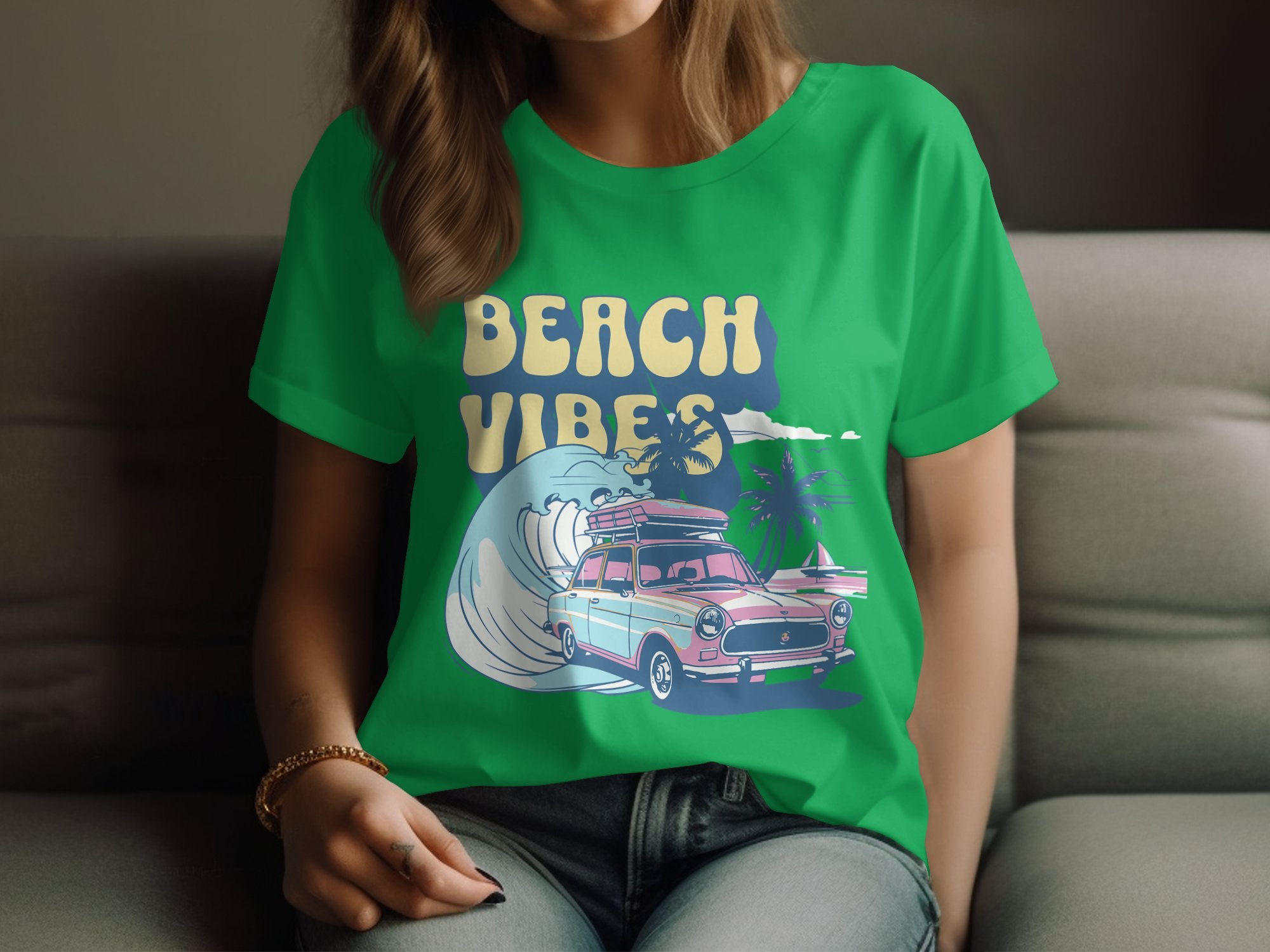 A person wearing a Garment Graphics green T-shirt is in full beach spirit, featuring a surf design with a vintage car, surfboards, palm trees, and a wave. They sit comfortably on a gray couch.