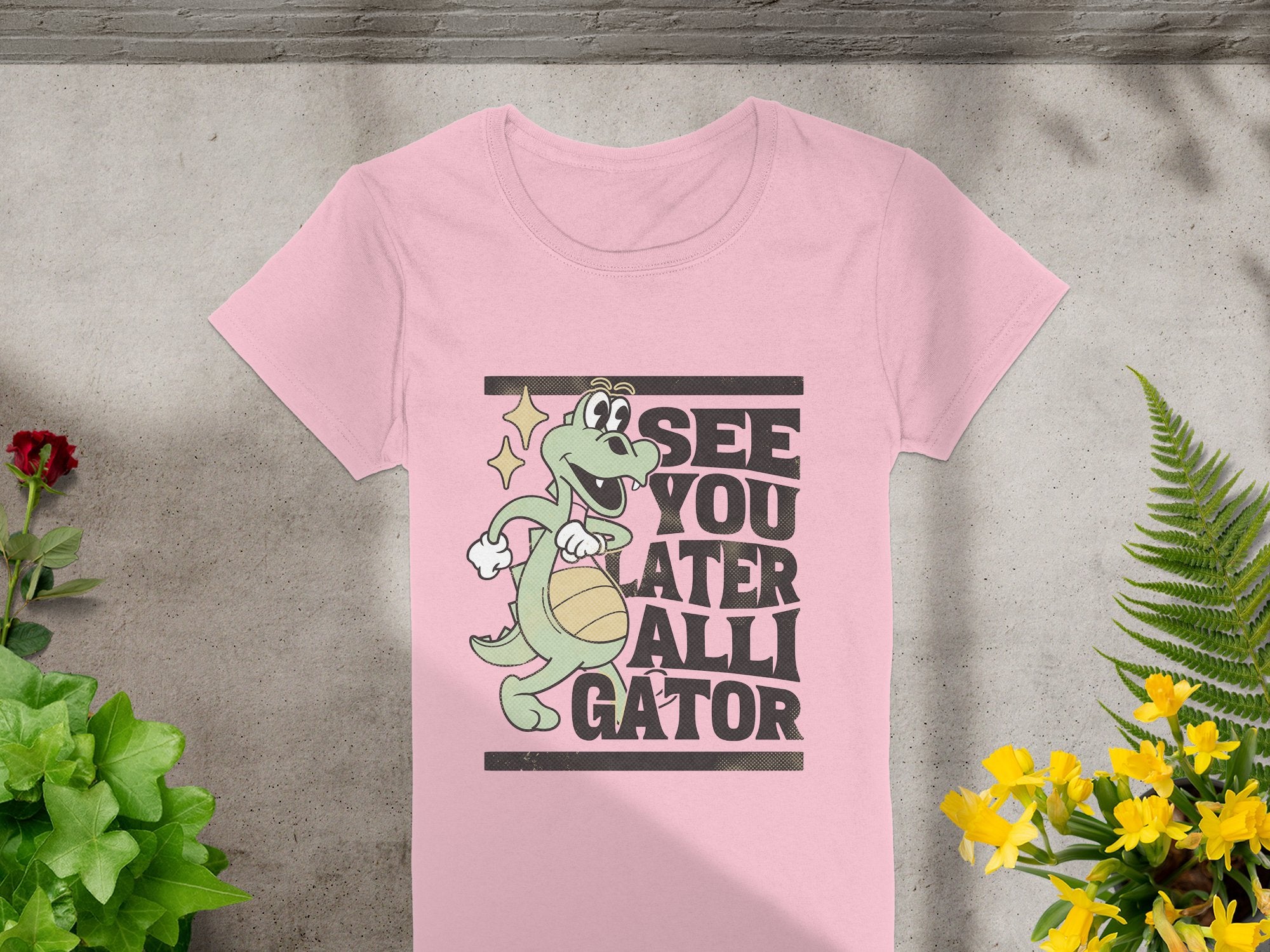 Garment Graphics presents the Alligator T-Shirt for women, featuring a whimsical cartoon of a smiling alligator with a basketball and the caption See you later, alligator, against a concrete backdrop with green plants and yellow flowers—ideal for humor lovers.