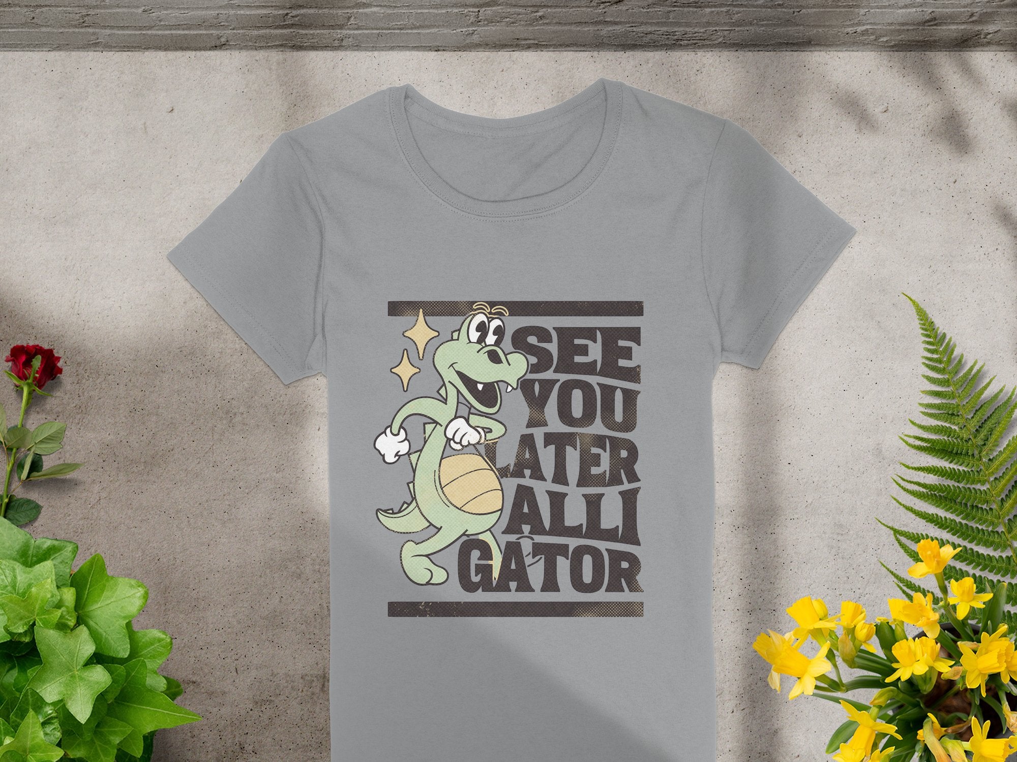 Garment Graphics offers a womens gray t-shirt featuring a cartoon alligator with a basketball and the phrase See you later alligator, set on concrete with greenery, red flowers on the left, and yellow on the right.