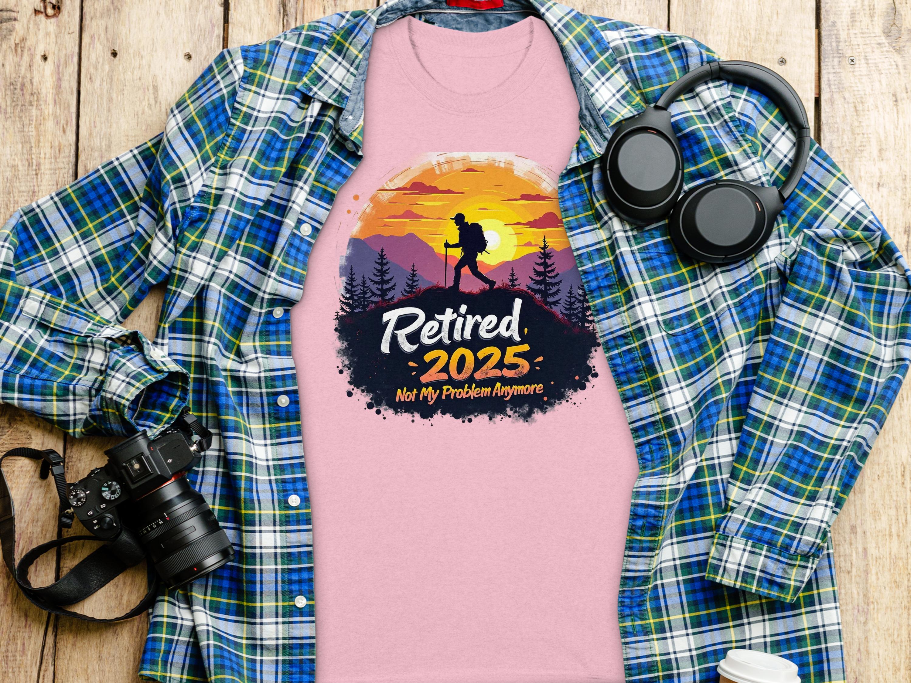 A folded pink Garment Graphics T-shirt with Retired 2025, Not My Problem Anymore and a hiking design lies under a golden sunset glow, accompanied by a blue plaid shirt, black headphones, a camera, and a coffee cup on the wooden surface.
