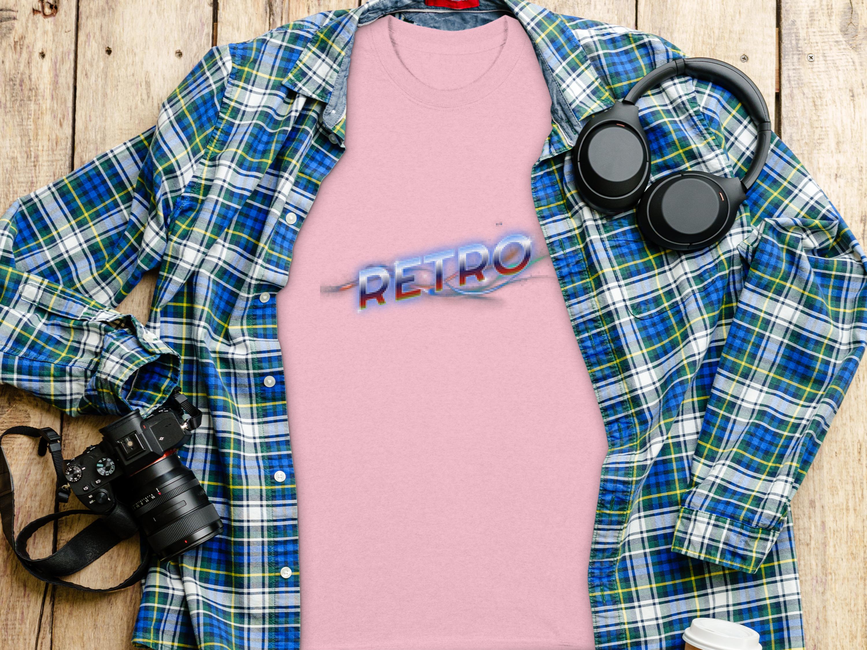 A pink Garment Graphics T-shirt with RETRO in blue and red text exuding vintage charm is displayed on a wooden surface, accompanied by a blue and green plaid shirt, camera, headphones, and a coffee cup lid.