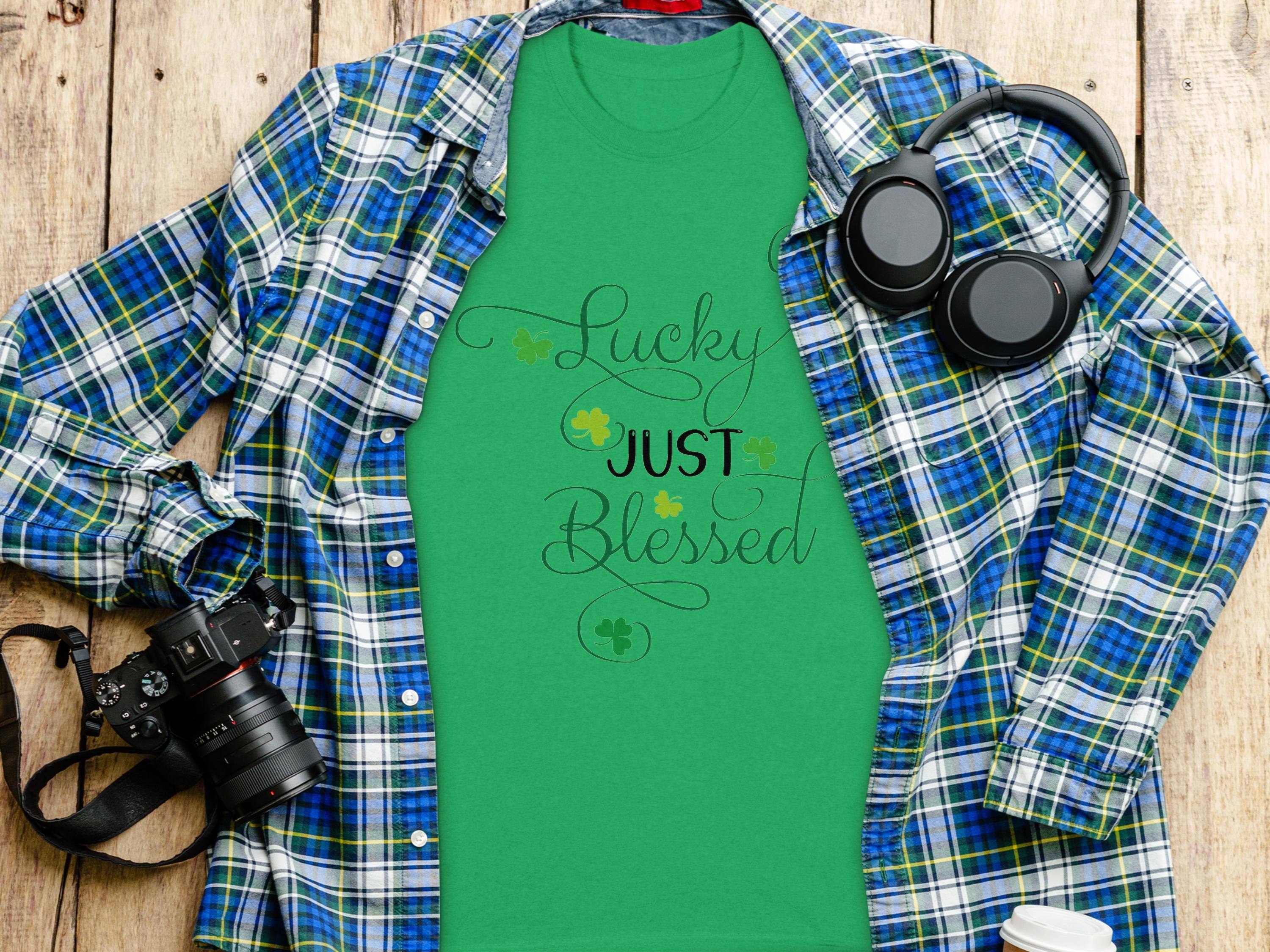 A classic fit green t-shirt by Royal Mail - 24 Tracked, featuring Lucky Just Blessed Shamrock and shamrock designs, rests on a checkered shirt. Nearby are black headphones, a camera, and a to-go coffee cup on the wooden surface.
