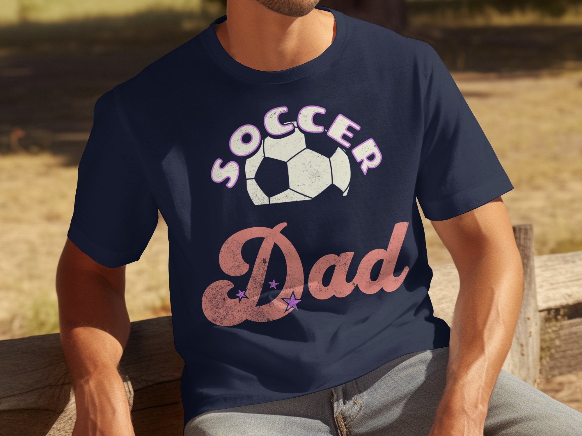 A dad sits outdoors on a wooden bench in a Garment Graphics navy blue T-shirt, featuring Soccer Dad with a soccer ball. The backdrop is a grassy landscape.