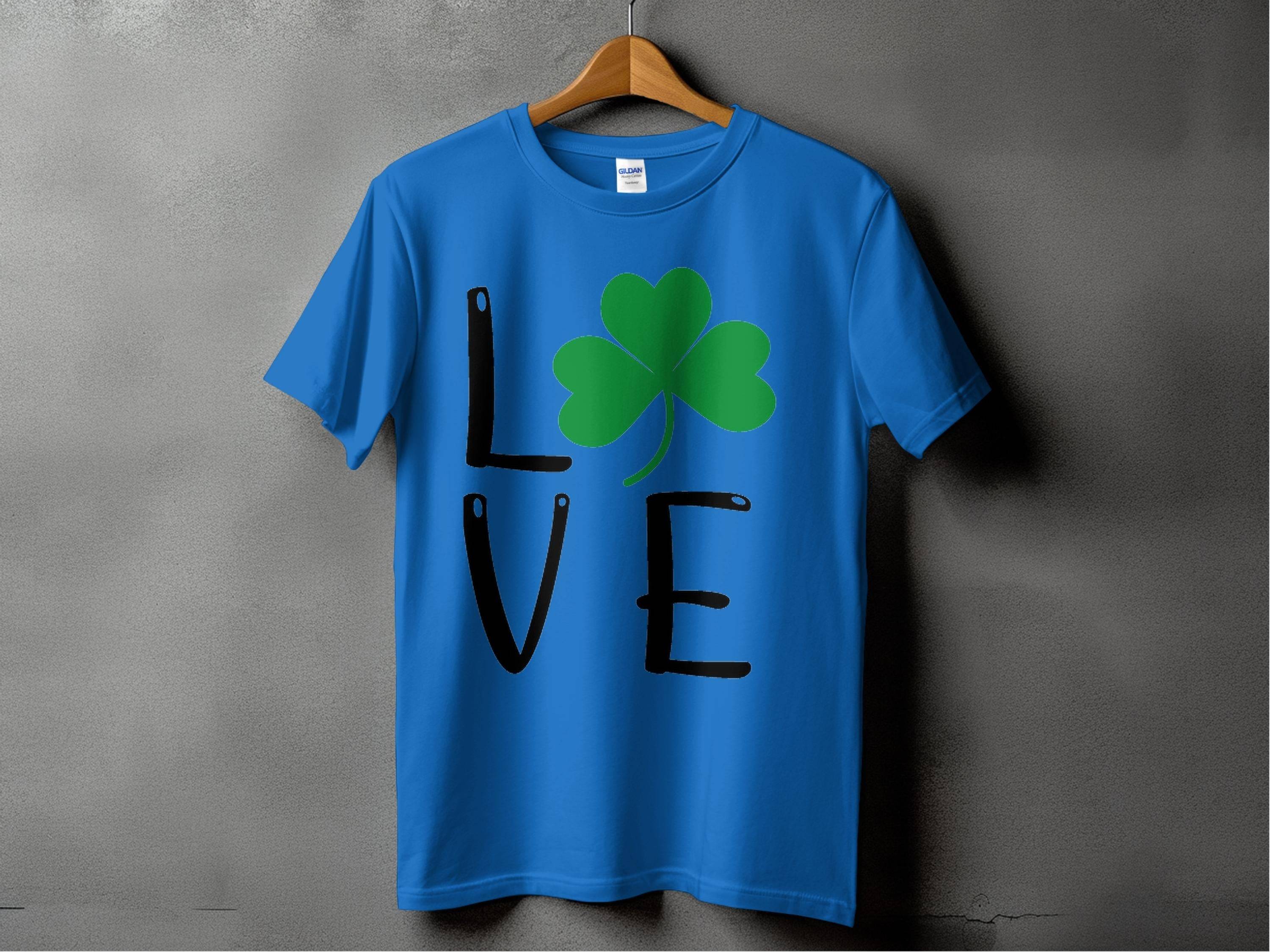 On a gray backdrop, a blue Garment Graphics t-shirt displays the word LOVE, featuring a green clover replacing the O, capturing Irish essence.