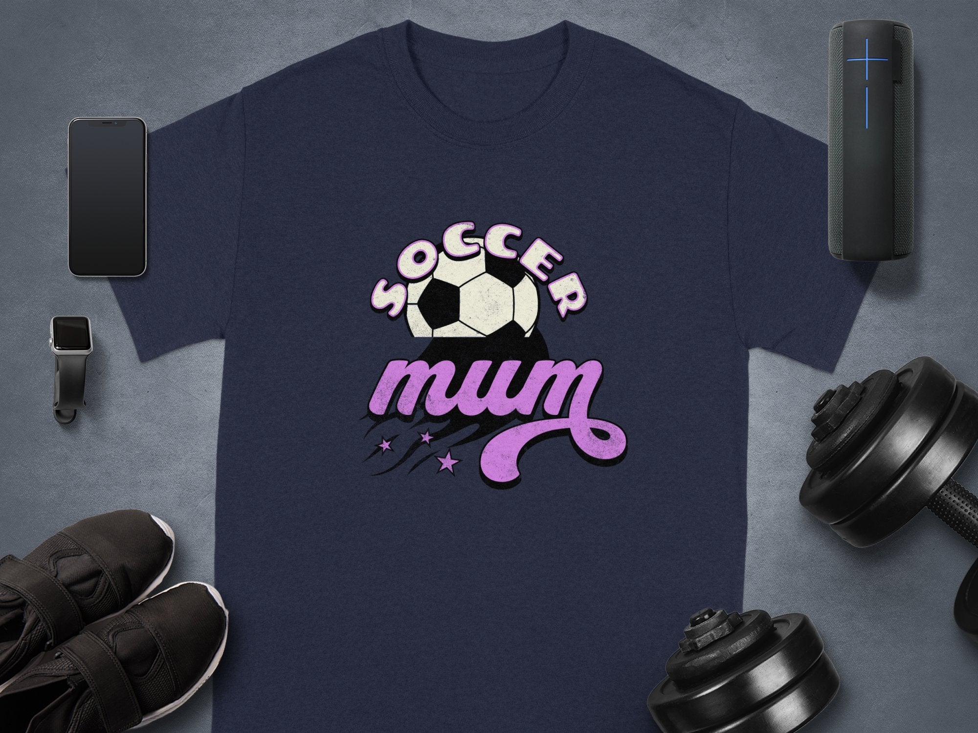 A navy graphic T-shirt by Garment Graphics, showcasing Soccer Mum in white and pink with a soccer ball, lies alongside a smartwatch, smartphone, wireless speaker, sneakers, and dumbbells on a gray surface.