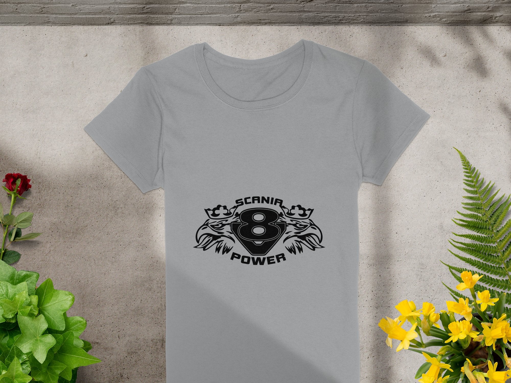 A gray T-shirt by Garment Graphics displays a central black design with SCANIA 8 POWER amid stylized flames or eagle heads. It is placed flat on concrete, decorated with surrounding plants and flowers.