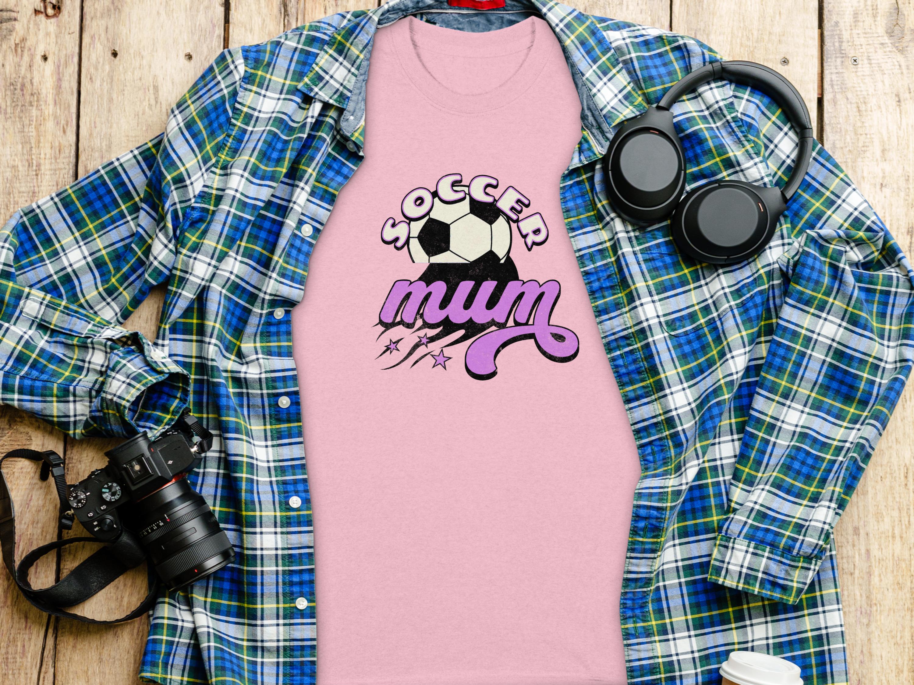 A flat lay showcases Garment Graphics classic fit Soccer Mum pink T-shirt with a soccer ball design, beneath an open blue and green plaid shirt. Nearby are headphones, a camera, and a disposable coffee cup on the wooden surface.