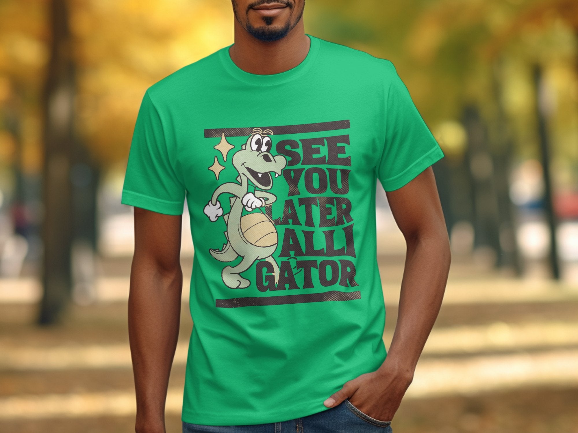 Dressed in a Garment Graphics casual green t-shirt, featuring a cartoon alligator playfully holding a basketball and the phrase See You Later Alligator, someone strolls along a picturesque tree-lined path adorned with vibrant autumn foliage.