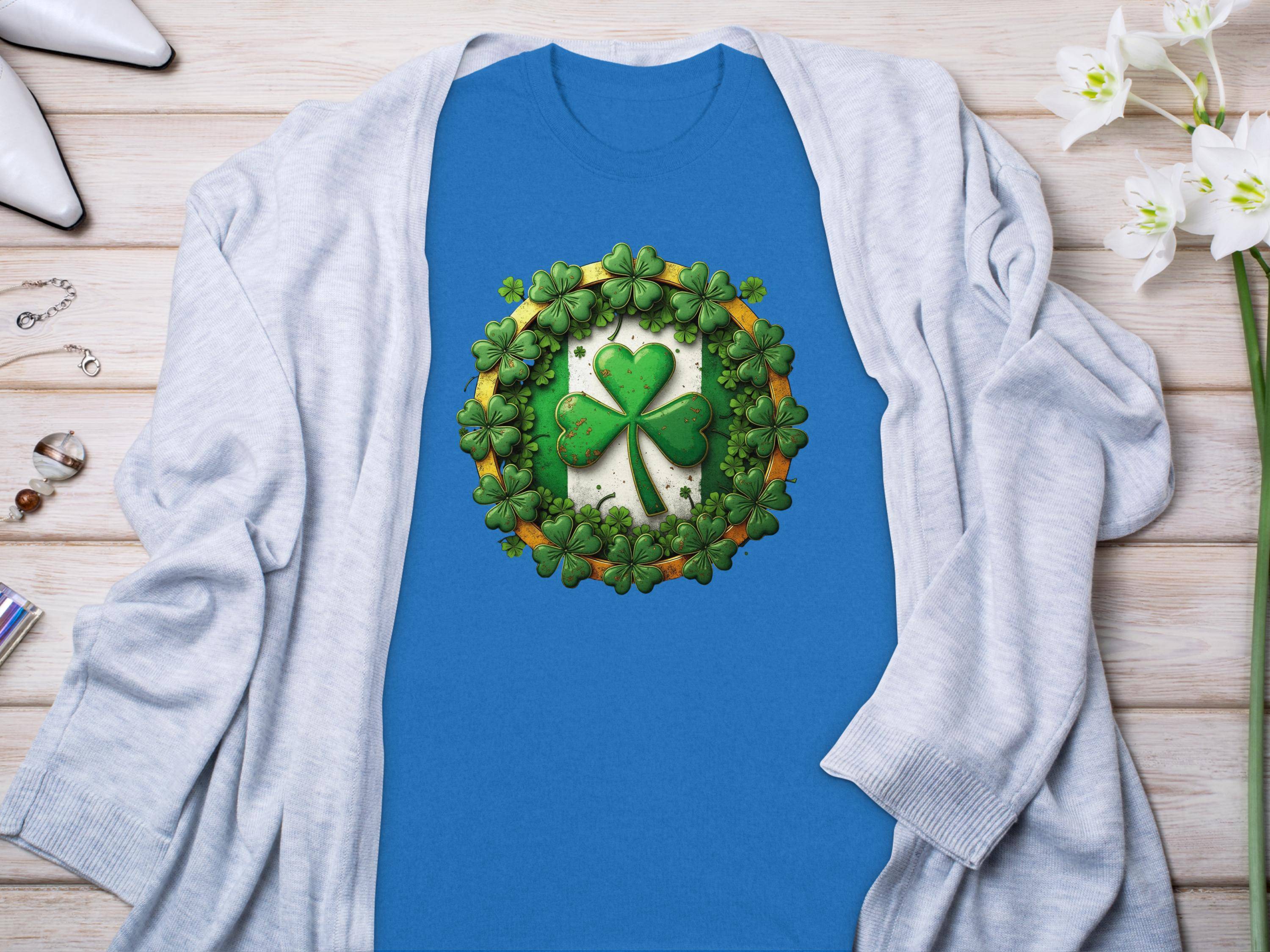 A Garment Graphics blue graphic T-shirt with a central shamrock clover and smaller shamrocks, paired with a light gray cardigan, creates a St. Patricks Day ensemble complemented by flowers, jewelry, and white shoes on a wooden surface.