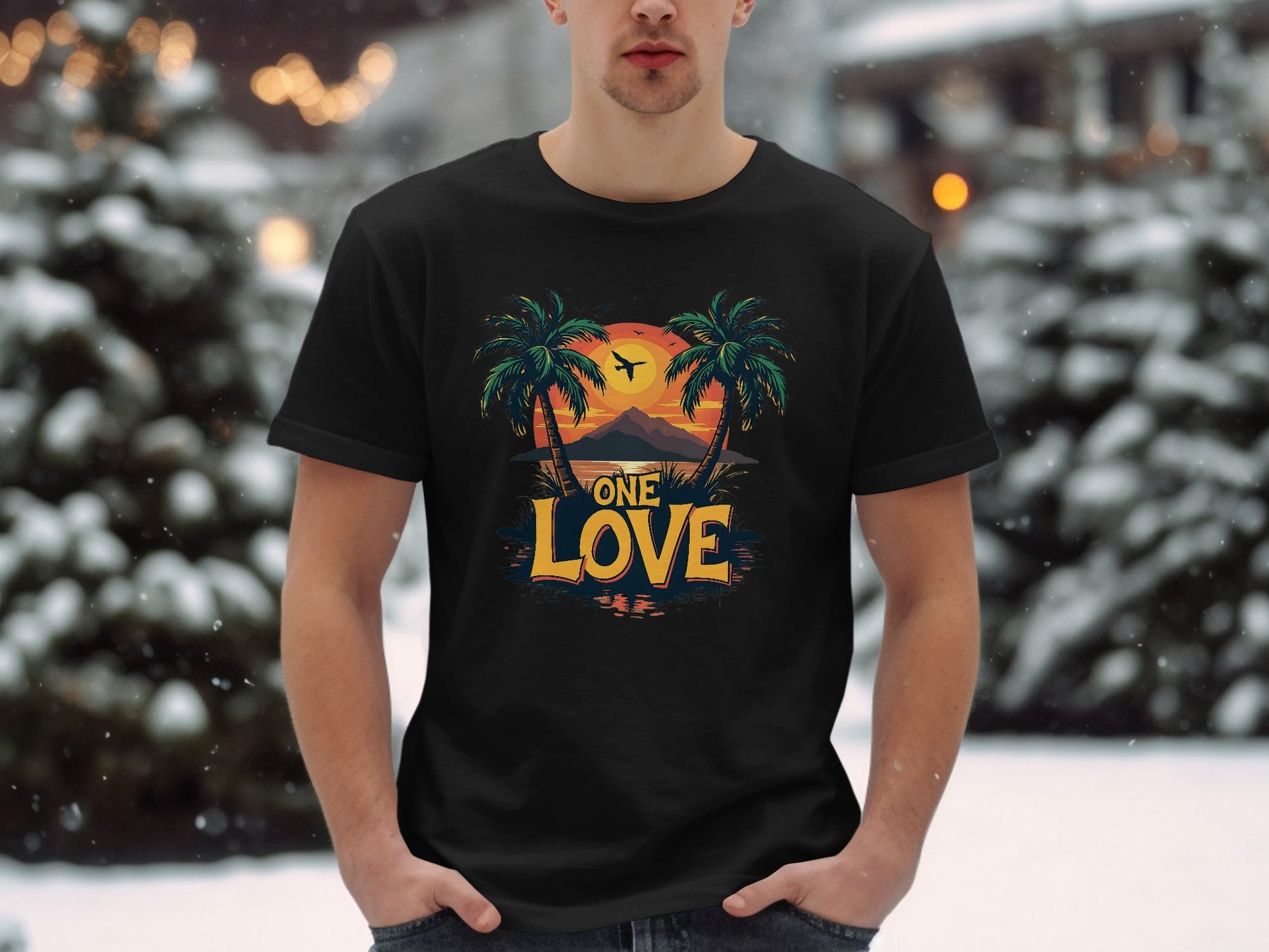 In a snowy setting, someone wears a black Garment Graphics T-shirt featuring palm trees, a tropical sunset, and a bird. ONE LOVE is boldly displayed below, bringing beach vibes to winter.