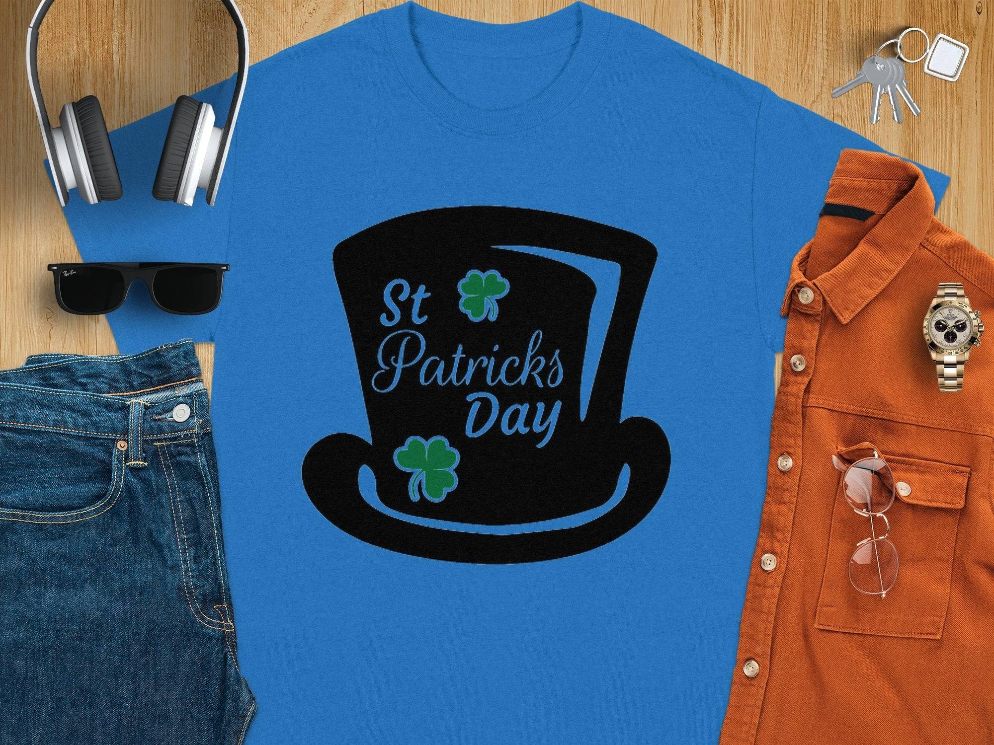 Flat lay of a Garment Graphics blue T-shirt with a black top hat and four-leaf clover design reading St Patricks Day, alongside denim jeans, rust-colored shirt, black sunglasses, headphones, keys, a wristwatch, and navy cap on wooden surface.