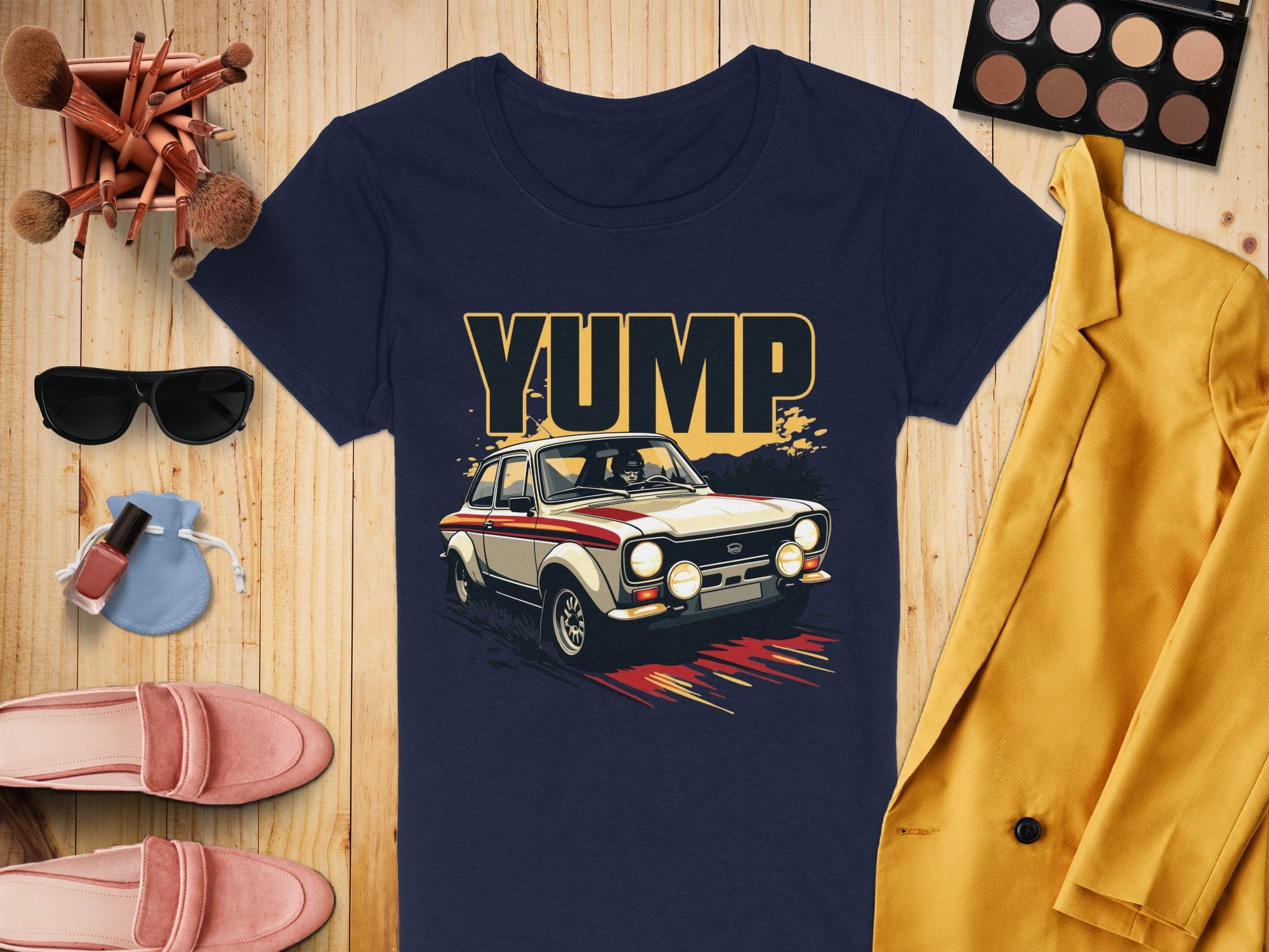 A navy Garment Graphics t-shirt with YUMP and a bold vintage racing car graphic is displayed on a wooden surface, surrounded by a yellow jacket, sunglasses, makeup brushes, an eyeshadow palette, pink shoes, perfume bottle, and nail polish.