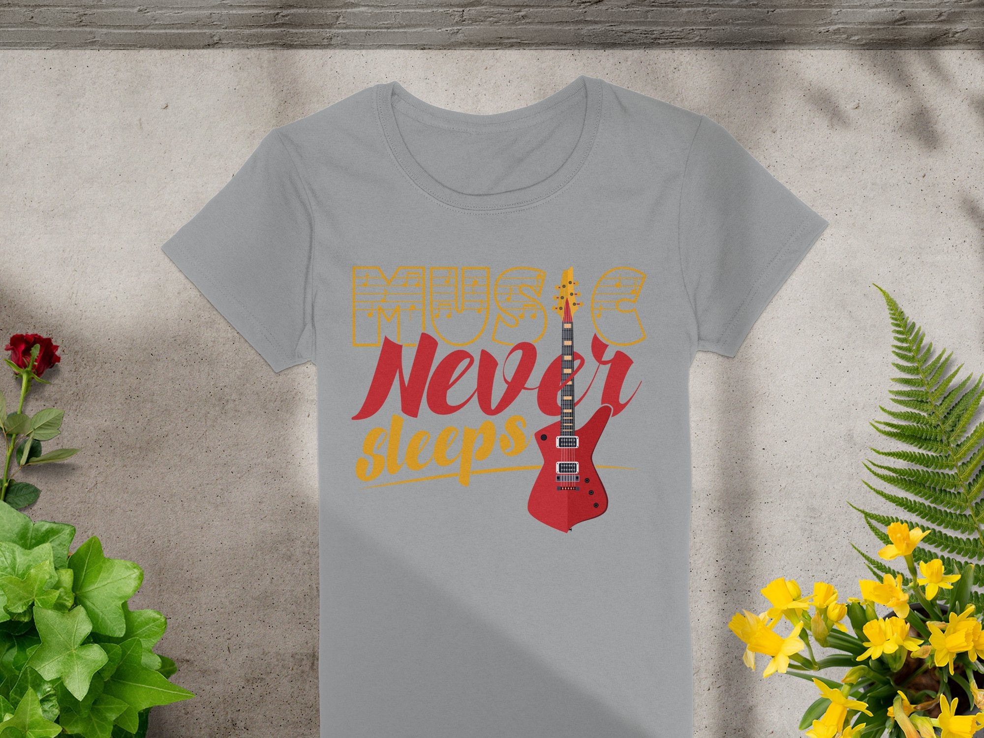 A Garment Graphics gray T-shirt with Music Never Sleeps in bold yellow and red letters and a striking red guitar motif lies surrounded by vibrant green plants and blooming yellow flowers.