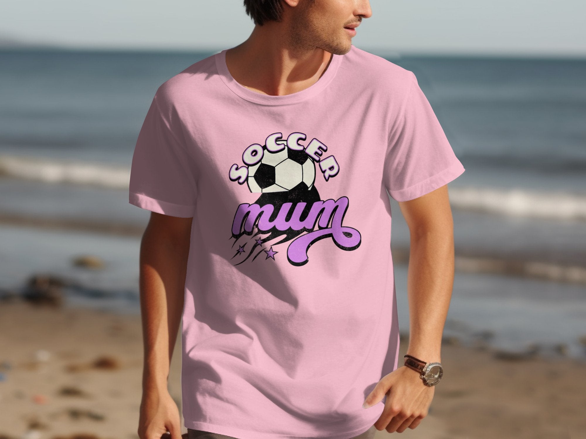 A person is walking on the beach in a light pink Garment Graphics classic fit tee, featuring a Soccer Mum slogan alongside a soccer ball design, with the ocean as a peaceful backdrop.