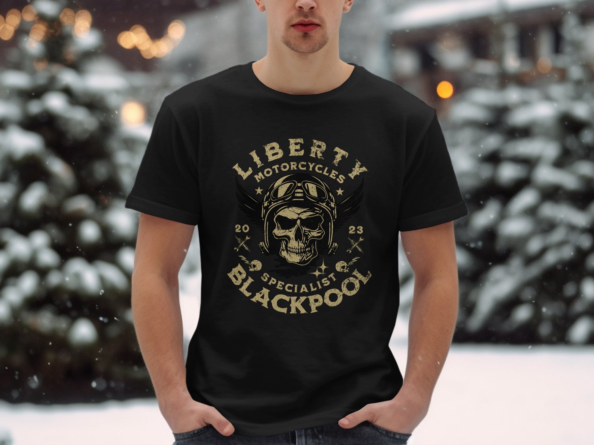 In the snowy outdoors, someone wears a Garment Graphics black T-shirt featuring a vintage skull with goggles and helmet design. The text says Liberty Motorcycles Specialist Blackpool 2023, embodying motorcycle culture. Snow-laden trees can be seen behind them.