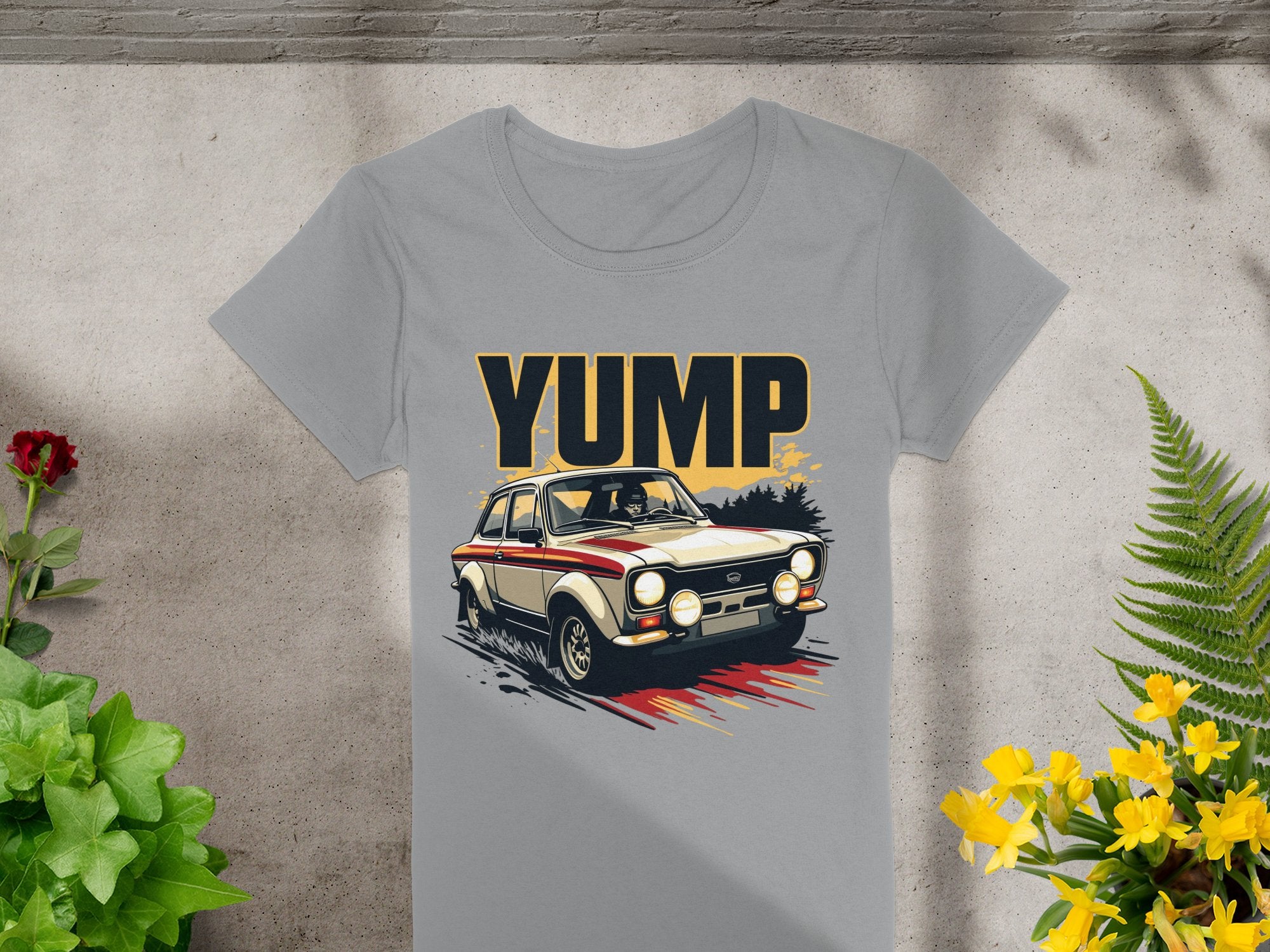 Garment Graphics presents a gray T-shirt with a vintage rally car graphic and YUMP text. Placed on concrete, its framed by vibrant green, red, and yellow plants for a retro look.