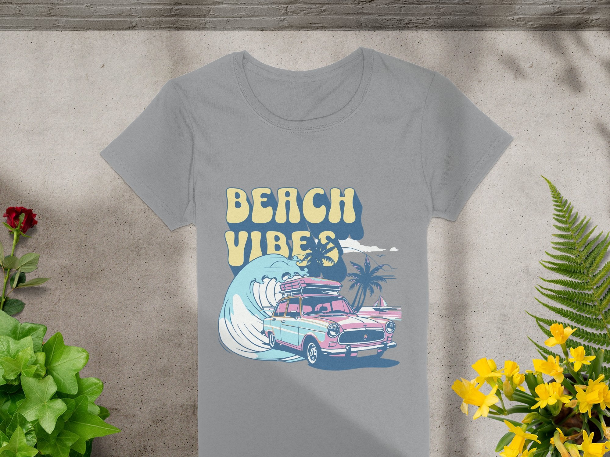 Garment Graphics gray T-shirt showcases a colorful vintage surf design with a car, surfboards, palm trees, and a large wave titled Beach Vibes, laid on gray fabric amid green, yellow, and red plants.