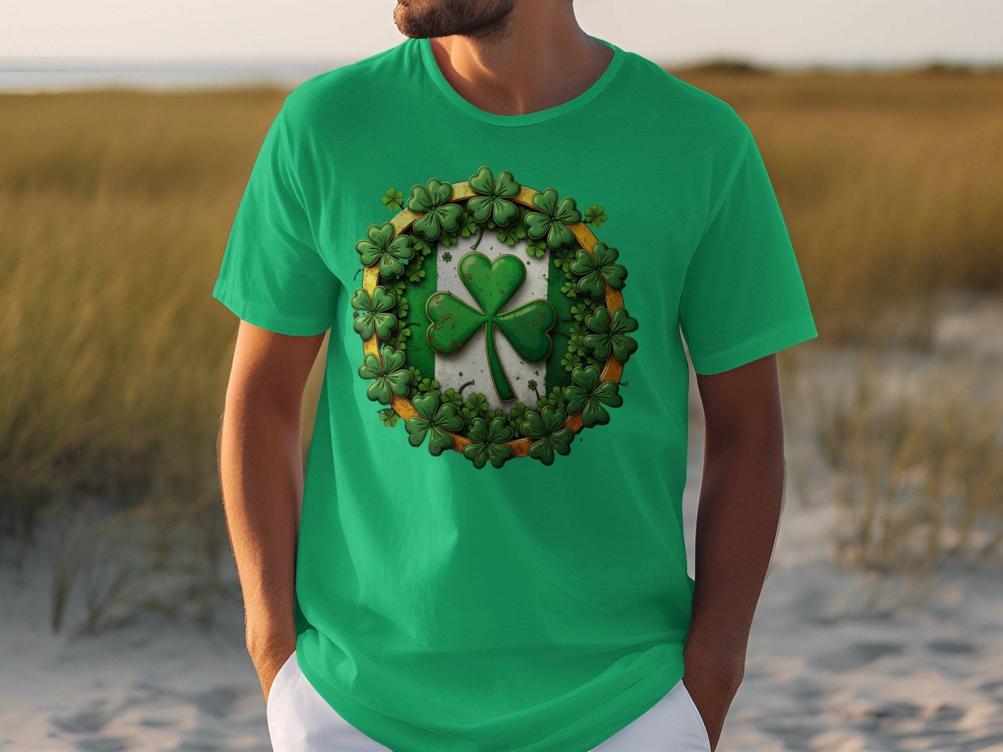 A person is wearing a Garment Graphics green T-shirt with a circular shamrock clover design and a larger central shamrock, suitable for St. Patricks Day, in an outdoor setting of grass and sand, hands in pockets.