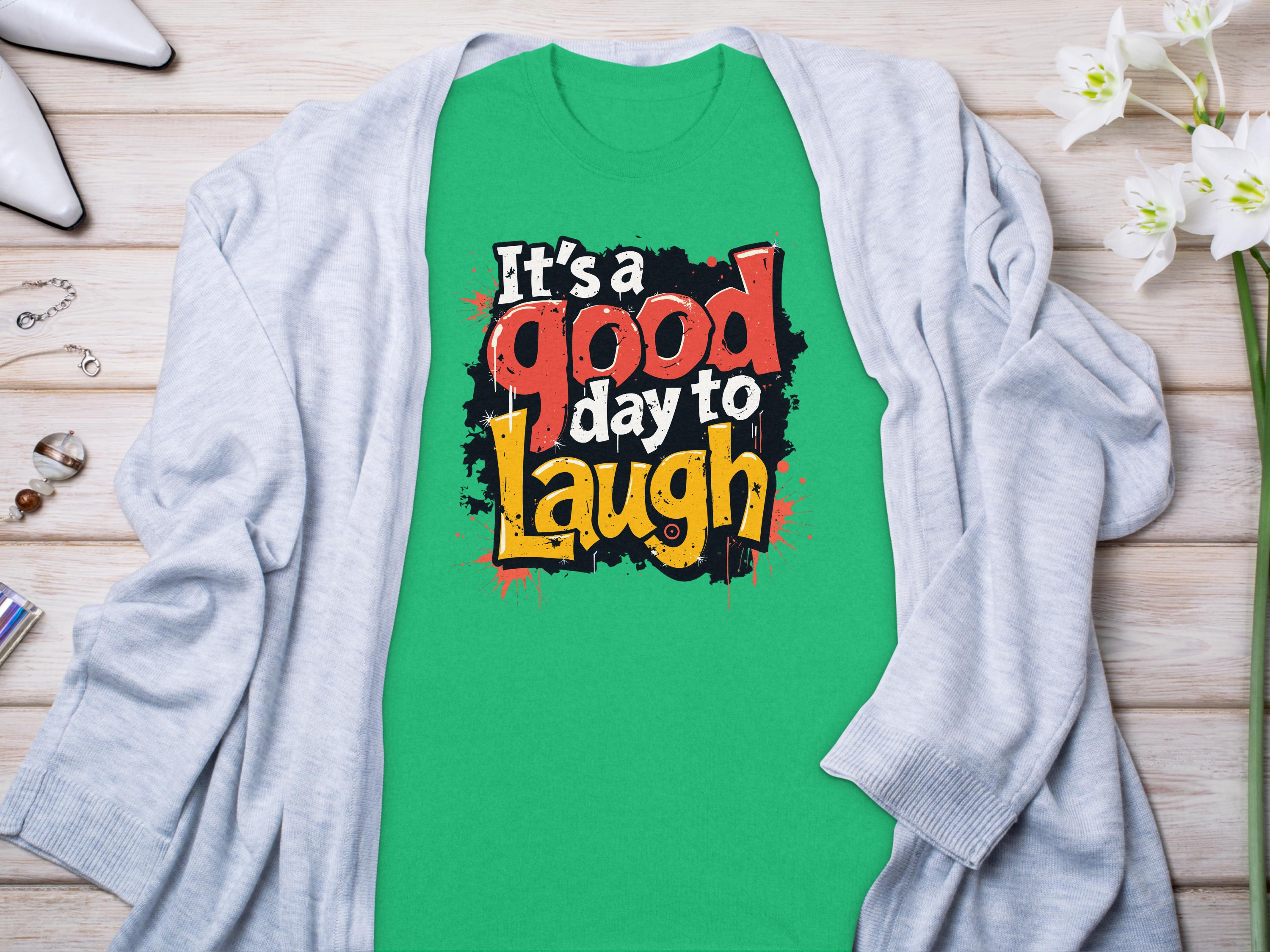 Garment Graphics Its a Good Day to Laugh t-shirt showcases vibrant colors and a cheerful message. Styled with a light gray cardigan, it lies on a wooden surface alongside white shoes, jewelry, and white flowers for an inviting look.