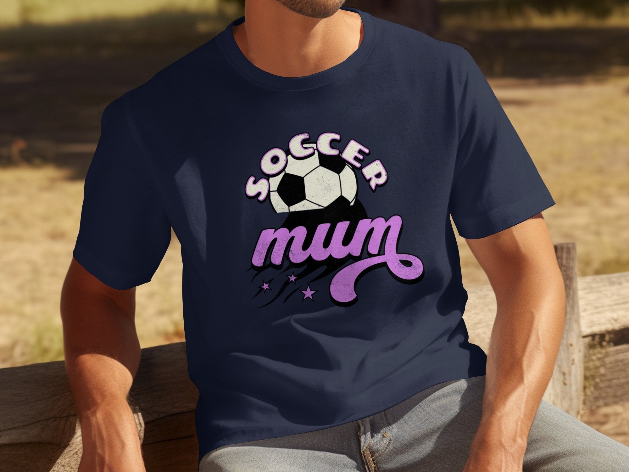 A person sits on a wooden bench wearing a classic fit dark blue T-shirt from Garment Graphics, featuring Soccer Mum and a soccer ball in white and pink. The green grass and dirt backdrop perfectly complement the sporty attire.
