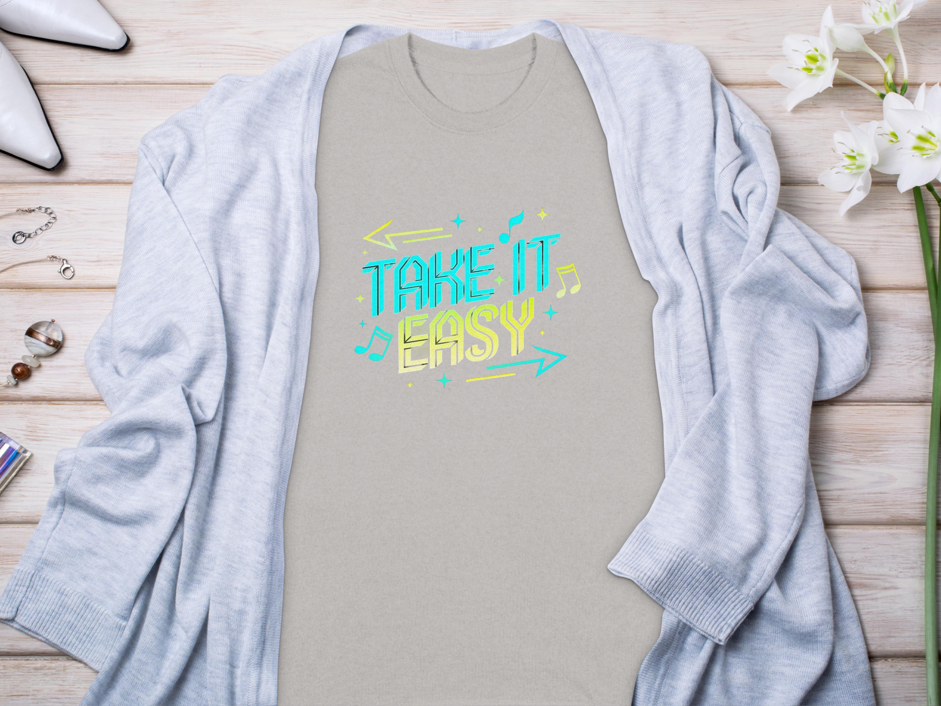 A Take It Easy shirt with colorful musical notes by Garment Graphics lies on a wooden surface, surrounded by a light gray cardigan, white flowers, jewelry, and high-heeled shoes.