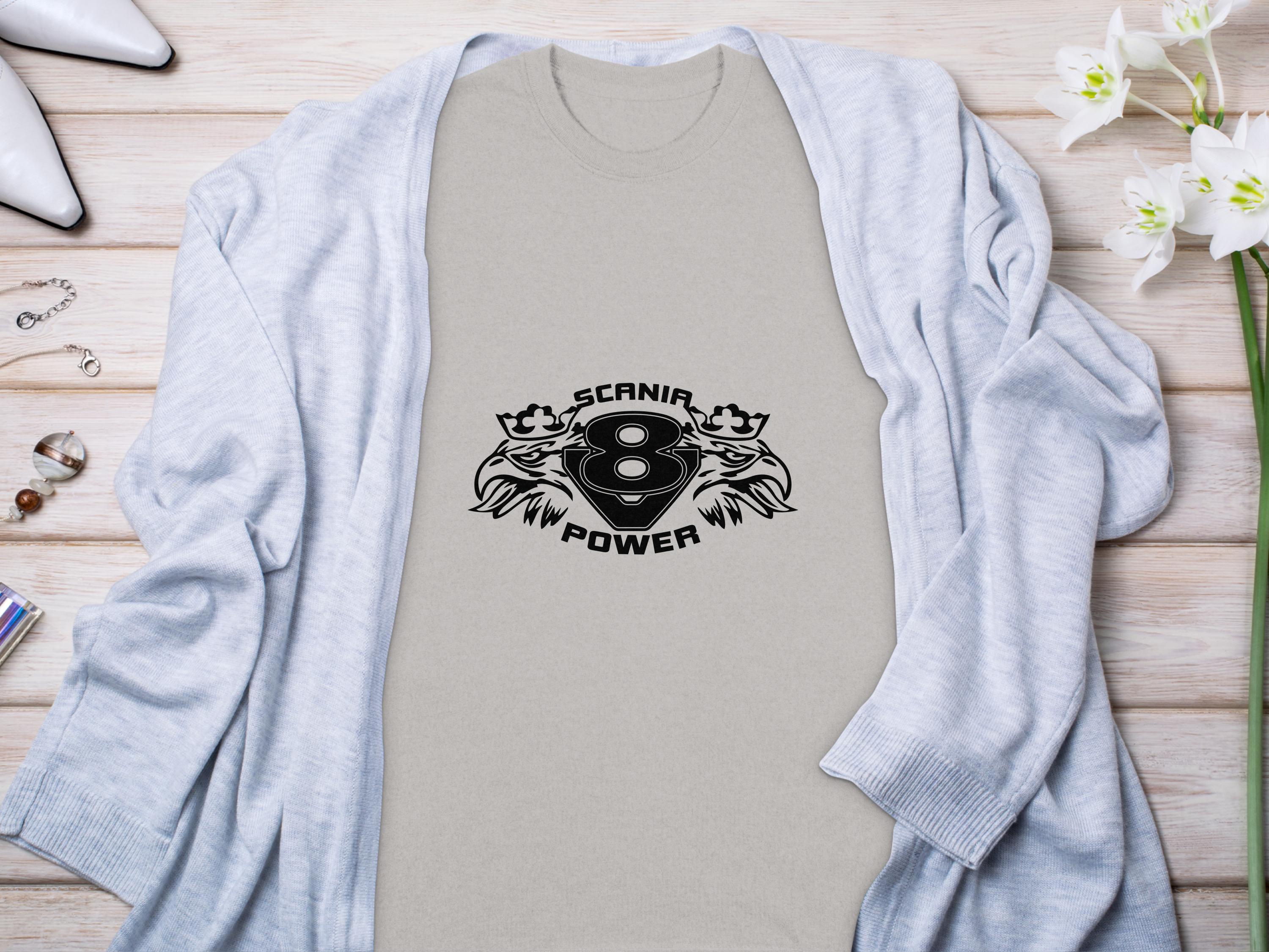 A Garment Graphics light T-shirt featuring a striking Scania Power Eagle design with two lions is laid on wood. Nearby are white heels, a gray sweater, and a vase of white flowers.