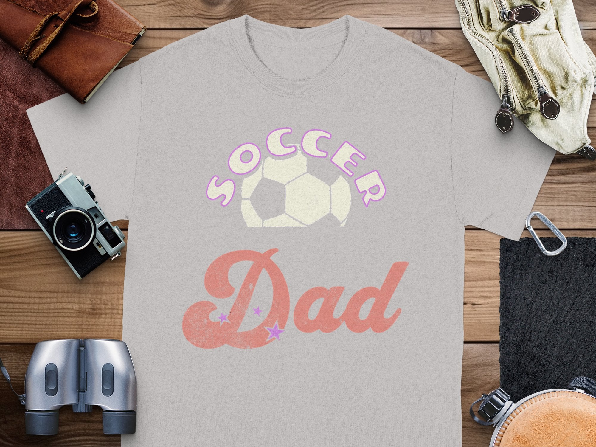 A Garment Graphics gray T-shirt with Soccer Dad in pink bubble letters above a soccer ball rests on wood, accompanied by a camera, binoculars, a leather pouch, and a wallet—ideal for the sporty dad who loves capturing every game moment.