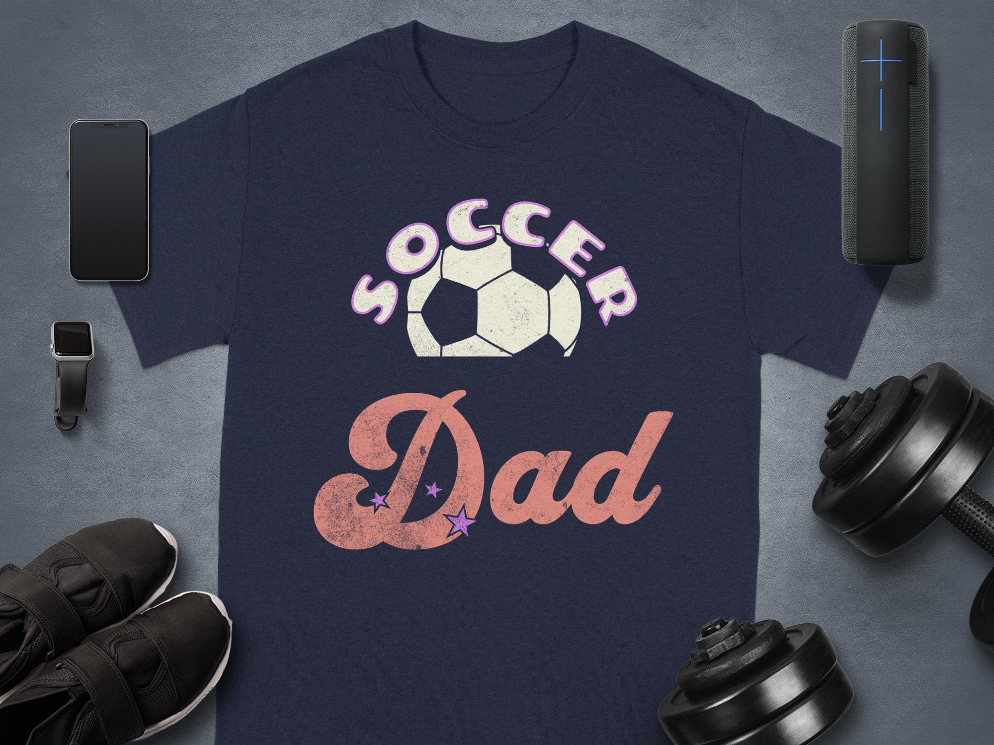 The Garment Graphics navy blue Soccer Dad T-shirt features a bold soccer ball graphic on a dark background, accompanied by a smartphone, smartwatch, wireless speaker, sleek black sneakers, and adjustable dumbbells.