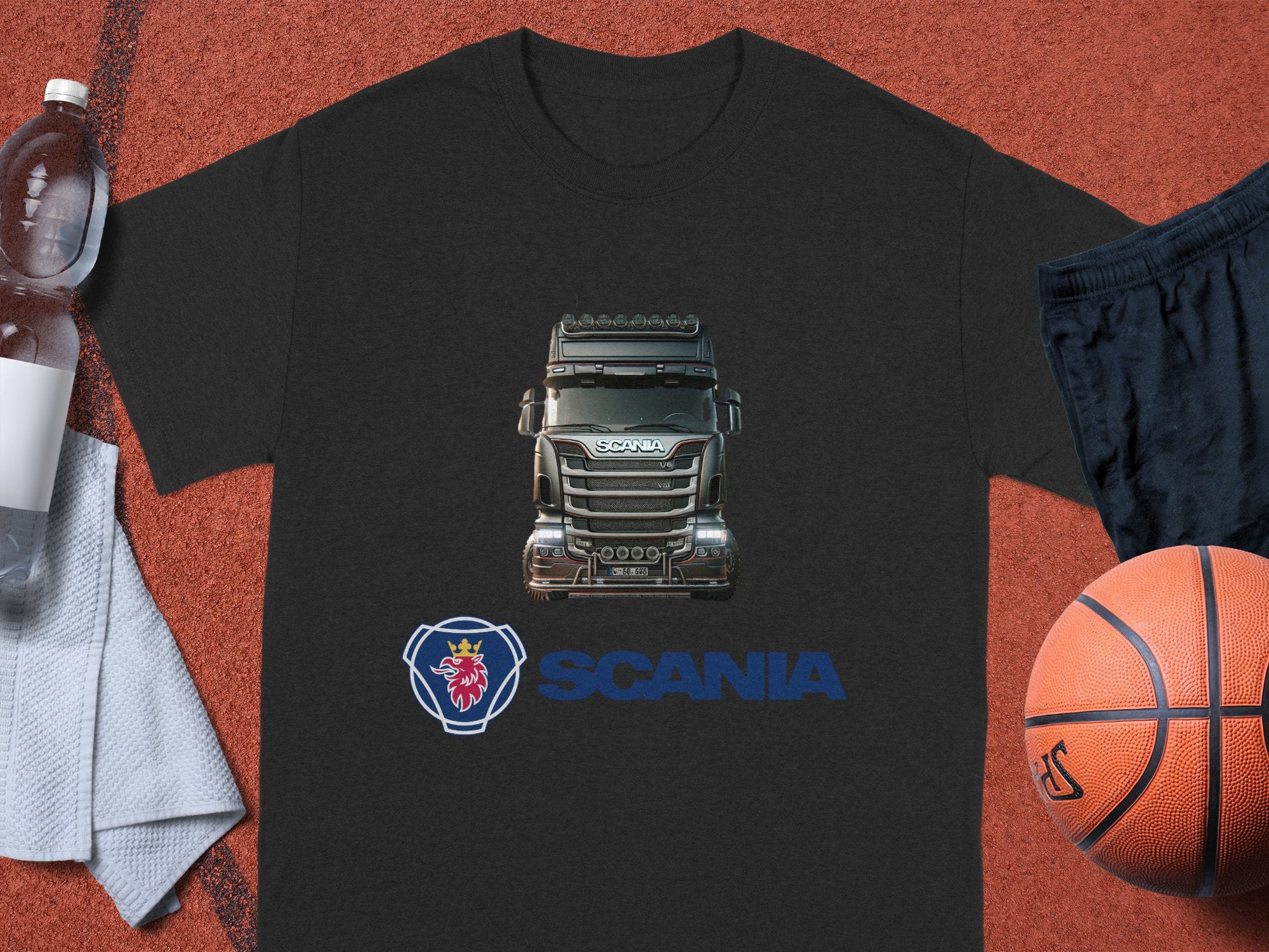 A black Garment Graphics T-shirt for truck enthusiasts, featuring a Scania truck image, SCANIA text, and a lion emblem on the front, is displayed on a textured brown surface with a basketball, black shorts, and a transparent water bottle nearby.