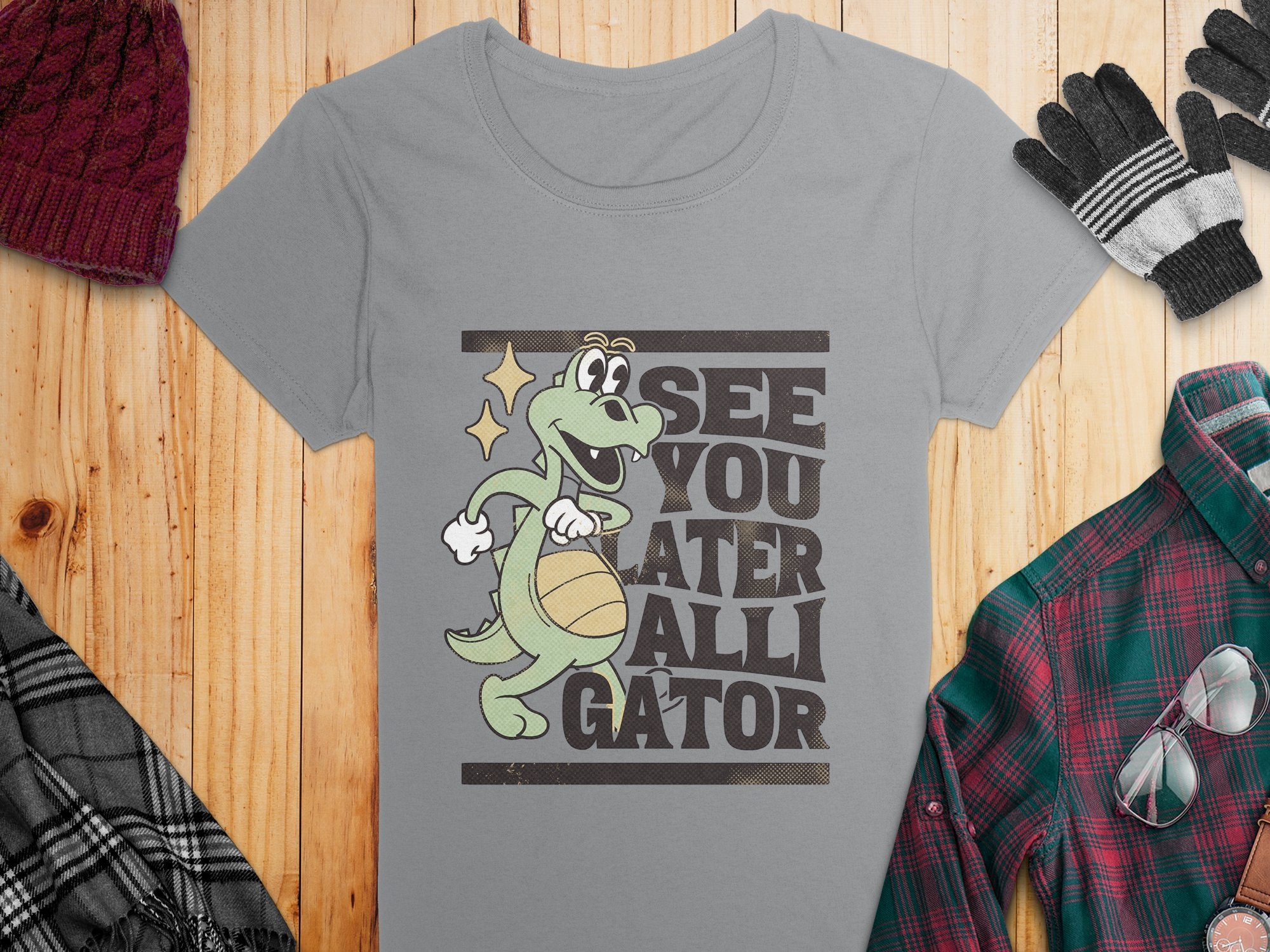 Garment Graphics Gray Alligator T-Shirt for women features a cartoon alligator holding a basketball with the phrase See you later alligator in bold. Styled with a red knit hat, gray gloves, plaid jacket, and glasses on a wooden surface.