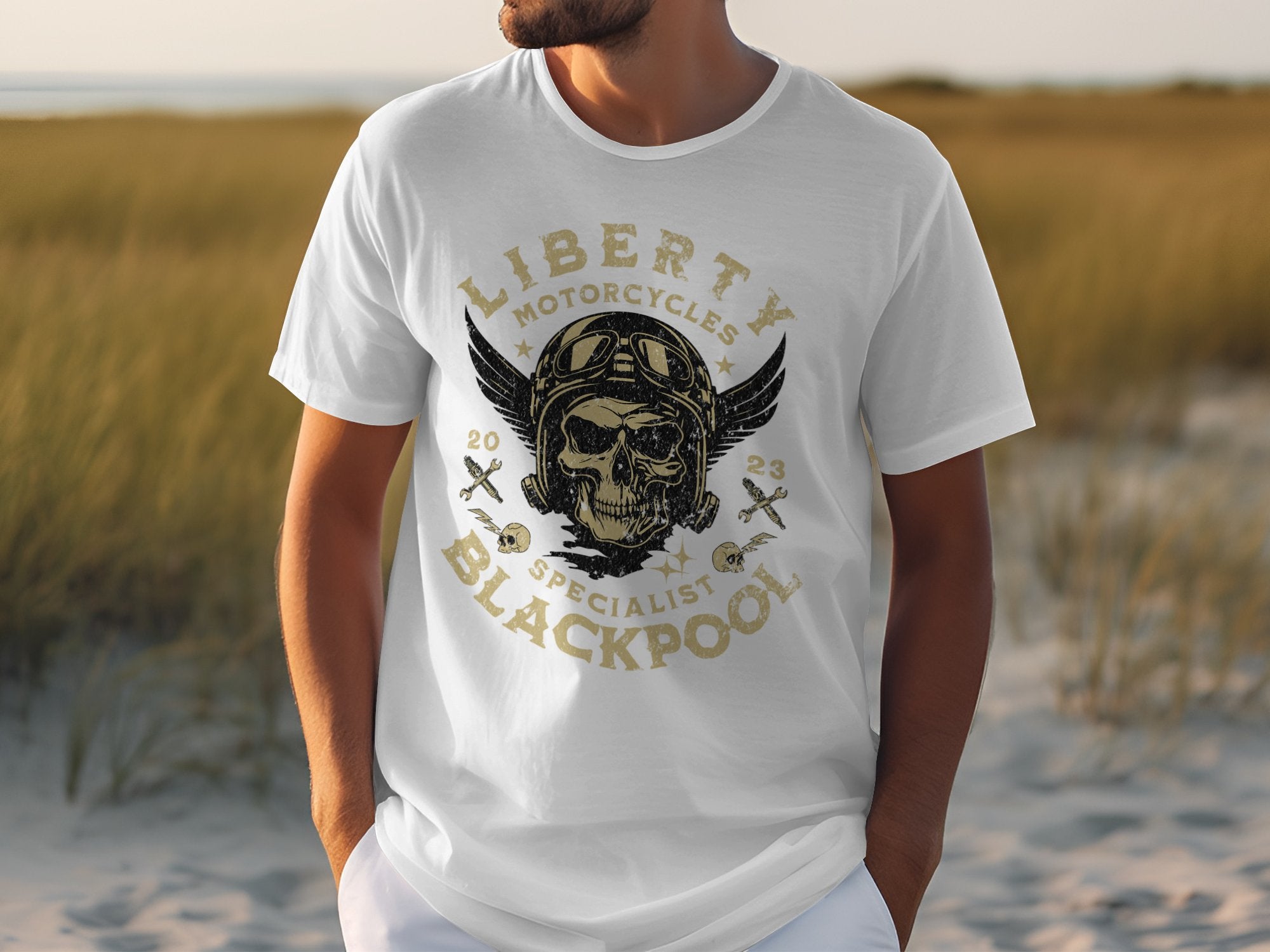 A man outside wears a Garment Graphics white T-shirt featuring a vintage skull with wings, helmet, and crossed tools design. The text Liberty Motorcycles Specialist Blackpool 2023 appeals to motorcycle enthusiasts. Theres grass meeting a sandy area in the background.