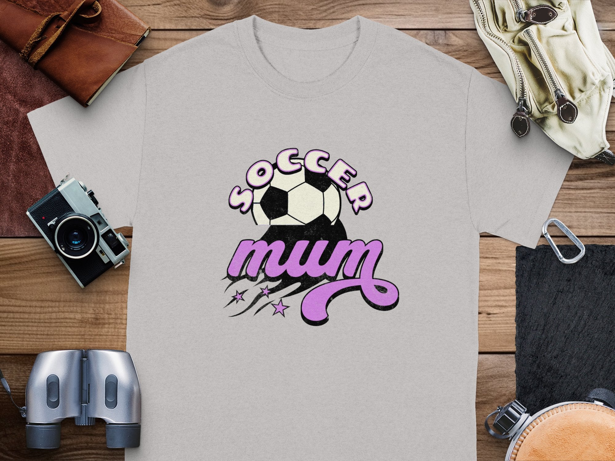 A light gray, classic fit T-shirt by Garment Graphics featuring a Soccer mum design with a soccer ball and pink accents is placed on wood. Nearby are a camera, binoculars, leather pouch, and gloves.