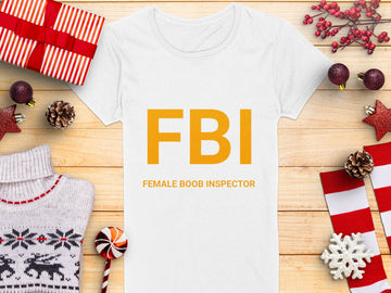 A white Garment Graphics T-shirt features yellow FBI Female Boob Inspector text, lying on wood. Its surrounded by holiday items: gifts, a deer-patterned sweater, pine cones, red ornaments, and a candy cane. Ideal as a gag gift and available in womens sizes.