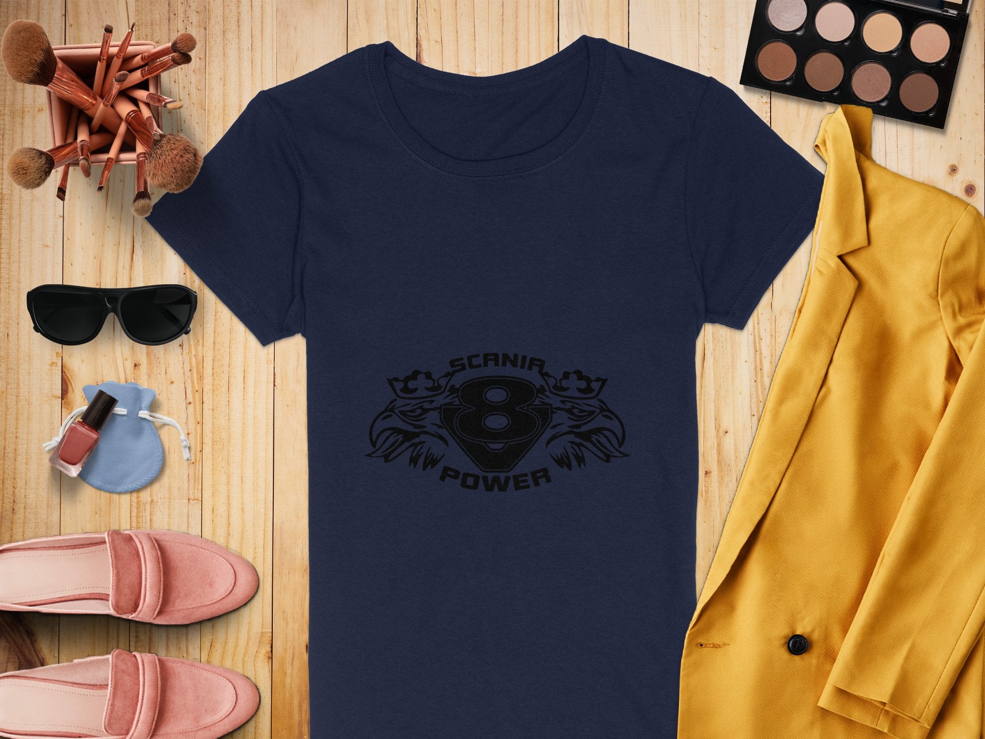 A flat lay image highlights a Garment Graphics navy T-shirt with Scania V8 Power design, paired with a yellow blazer, pink shoes, sunglasses, and a blue purse. Completing the look are makeup brushes, an eyeshadow palette with eagle heads, and nail polish on a wooden surface.