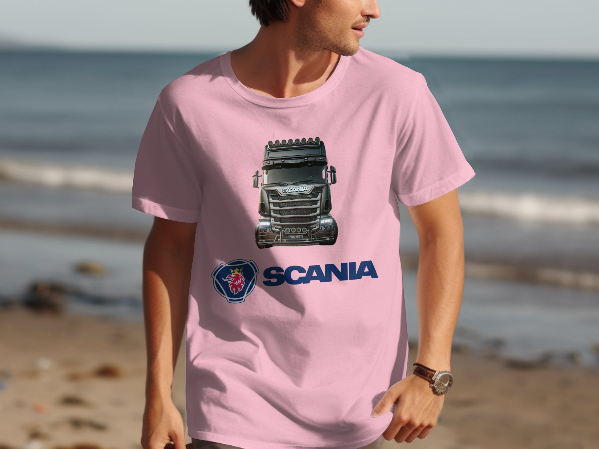 A truck enthusiast wears a pink Garment Graphics T-shirt featuring a Scania truck image and logo. They pose on a tranquil beach with the ocean in the background.