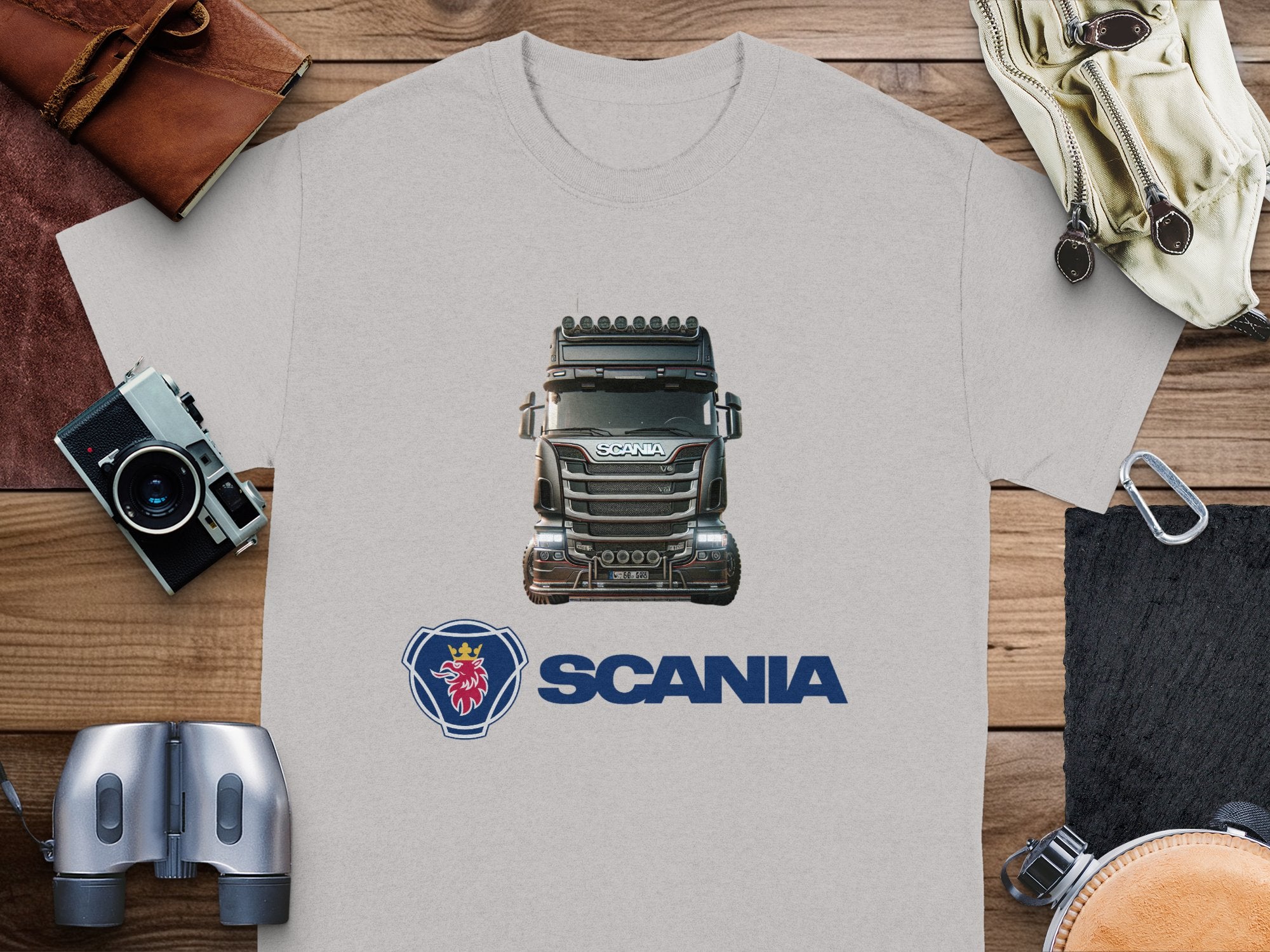 A Garment Graphics gray t-shirt, featuring a Scania truck and logo, is laid flat on a wooden surface. It’s perfect for truck enthusiasts. Nearby items include a brown leather pouch, camera, binoculars, pen, and a cream-colored watch in its case.
