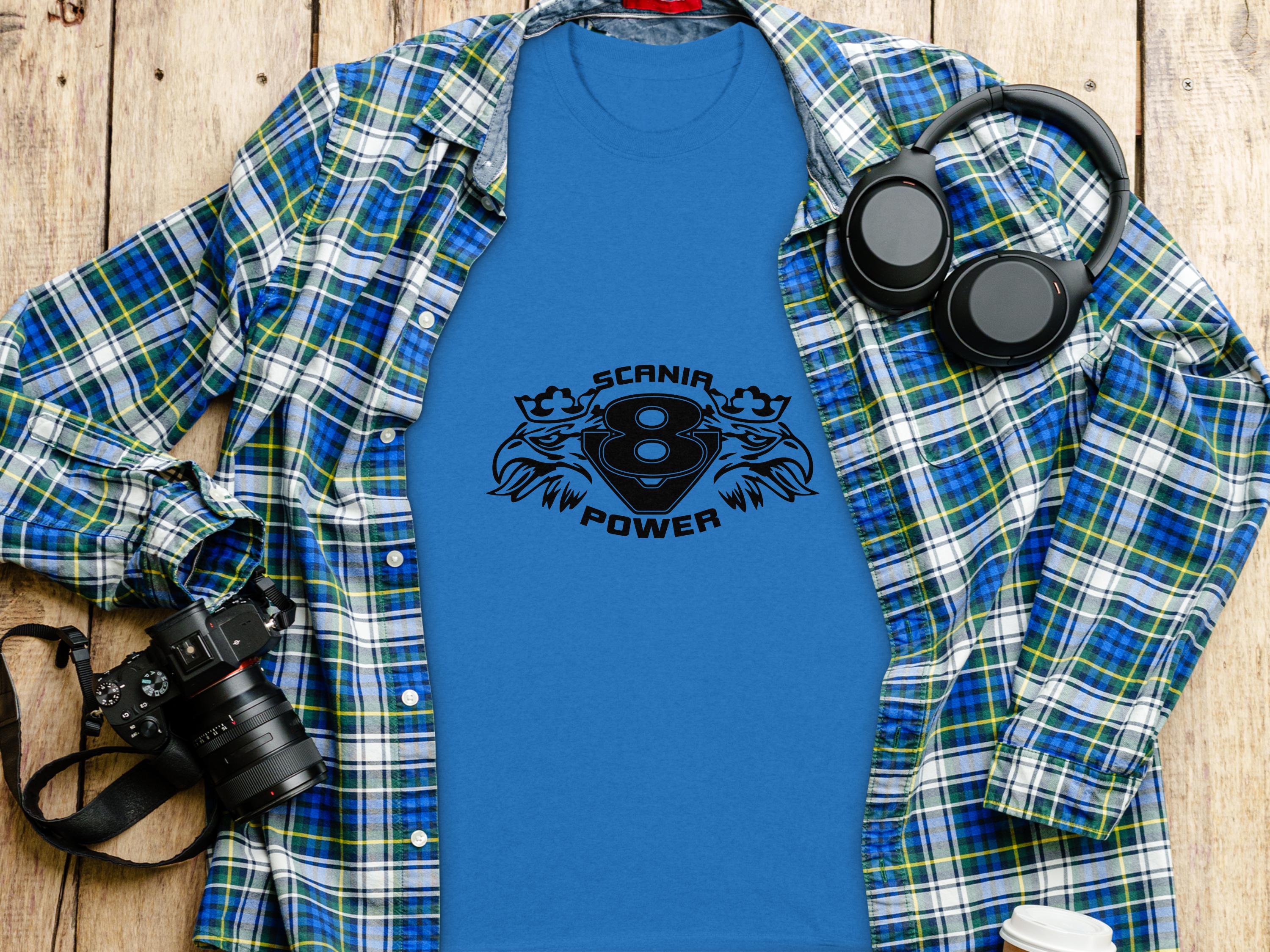 Flat lay of a blue graphic t-shirt featuring SCANIA POWER and an eagle logo by Garment Graphics, artfully arranged on a wooden surface. The stylish setup includes a blue plaid shirt, black headphones, camera, and coffee cup lid.