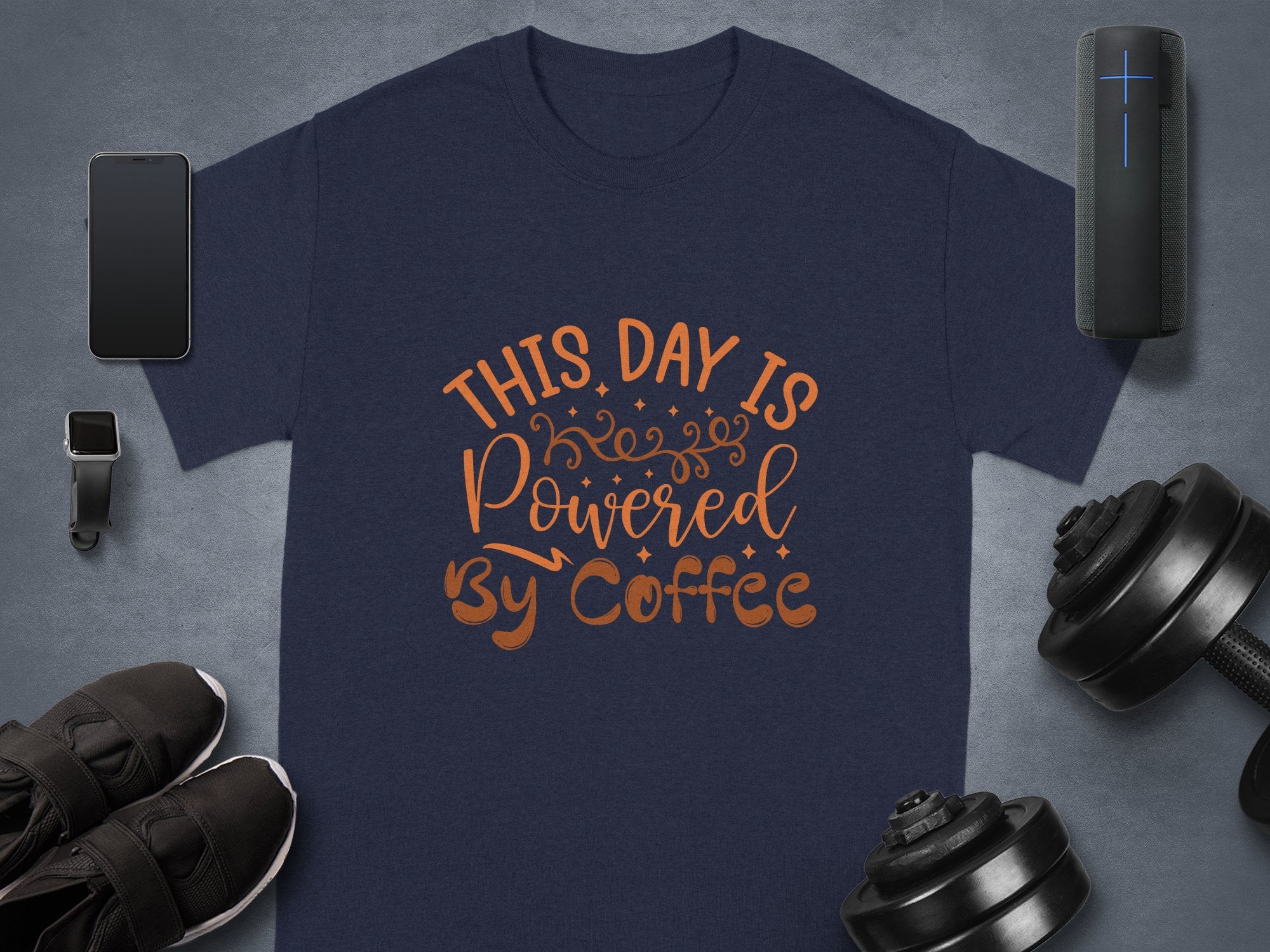 A flat lay photo features a Garment Graphics classic fit navy t-shirt with This Day is Powered by Coffee in brown. A smartphone, fitness tracker, wireless speaker, shoes, and dumbbell weights surround the coffee t-shirt on a gray background.
